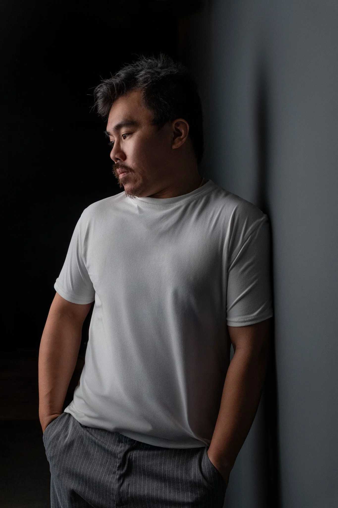 Man wearing a white T-shirt and gray pinstripe pants leaning against a wall, looking down with a contemplative expression.