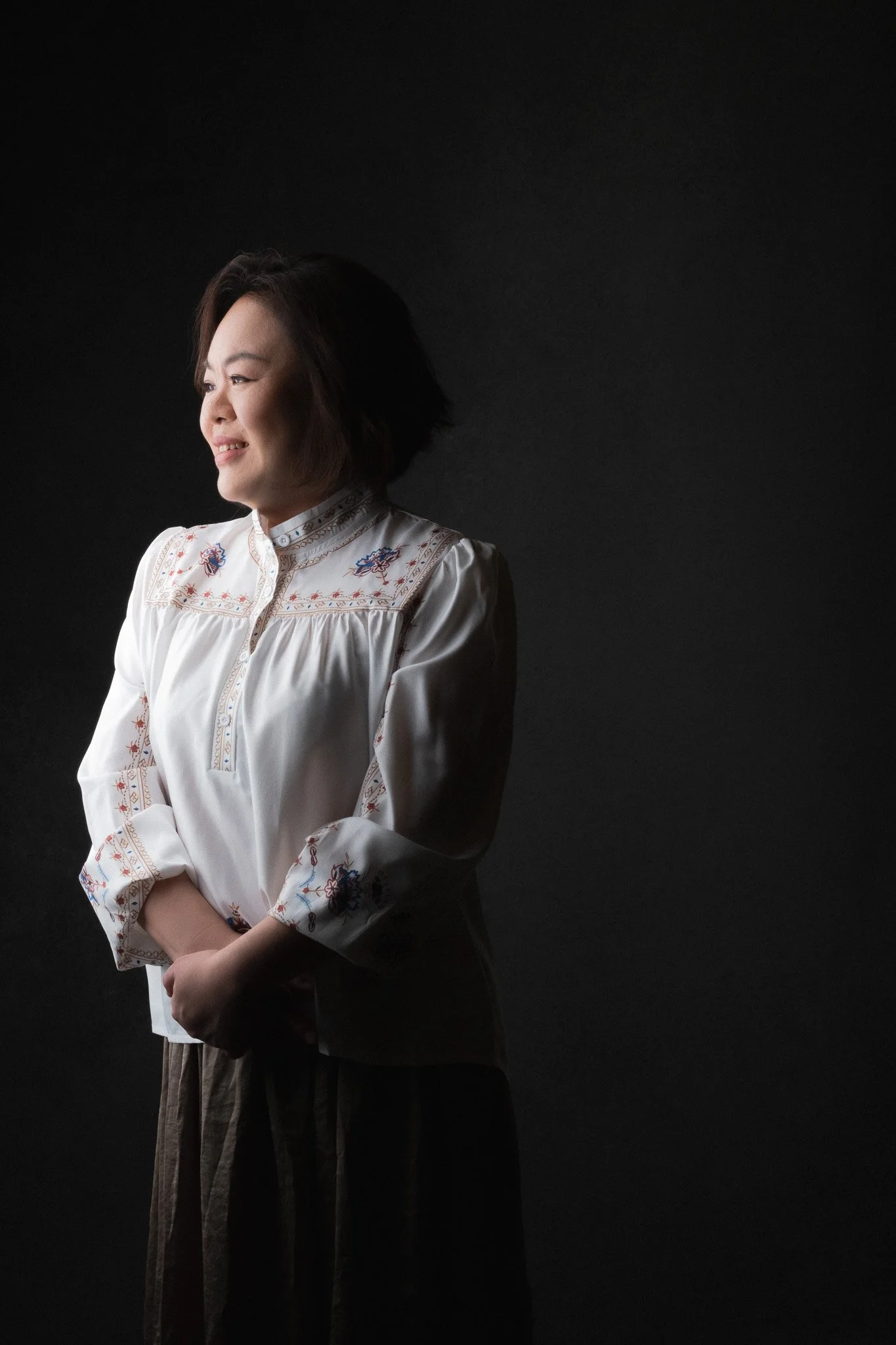 A woman with short dark hair smiling, wearing a white embroidered blouse, standing against a dark background.