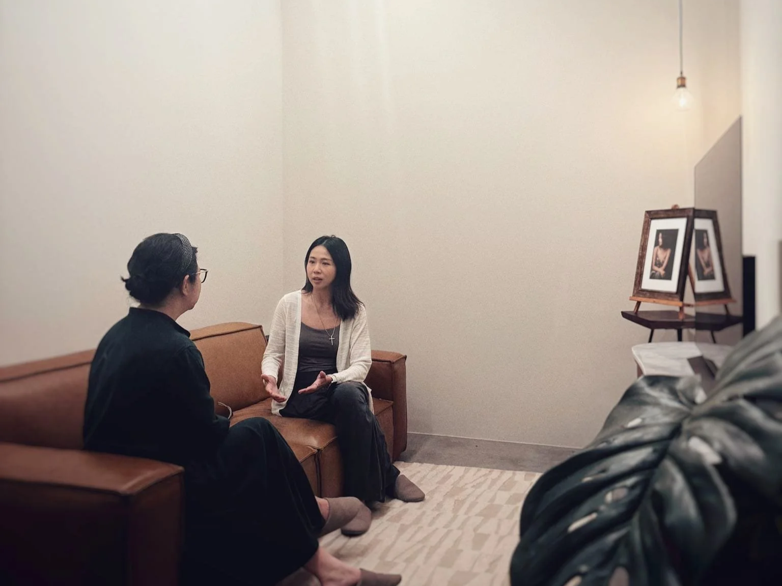 Two women are sitting on a brown sofa in a minimalistic room, engaged in a conversation. One woman has dark hair, wears a gray top, white cardigan, and a cross necklace. The other woman is wearing glasses, a black outfit, and has her hair pulled back. There are framed photographs on a stand to the right, and a small table with a book behind the sofa. A hanging light bulb illuminates the room.
