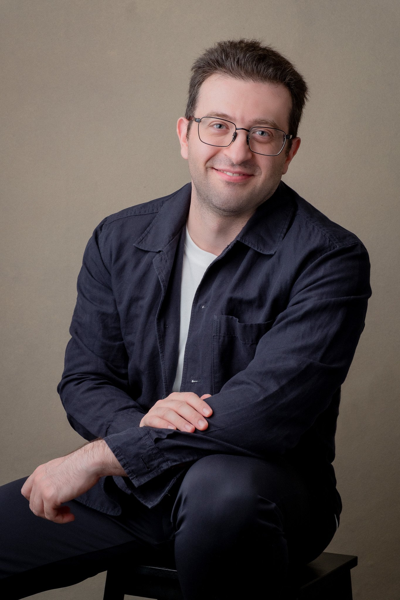 A man with short dark hair, wearing glasses, a dark blue shirt, and white undershirt, sitting on a black bench against a plain beige background, smiling and looking at the camera.