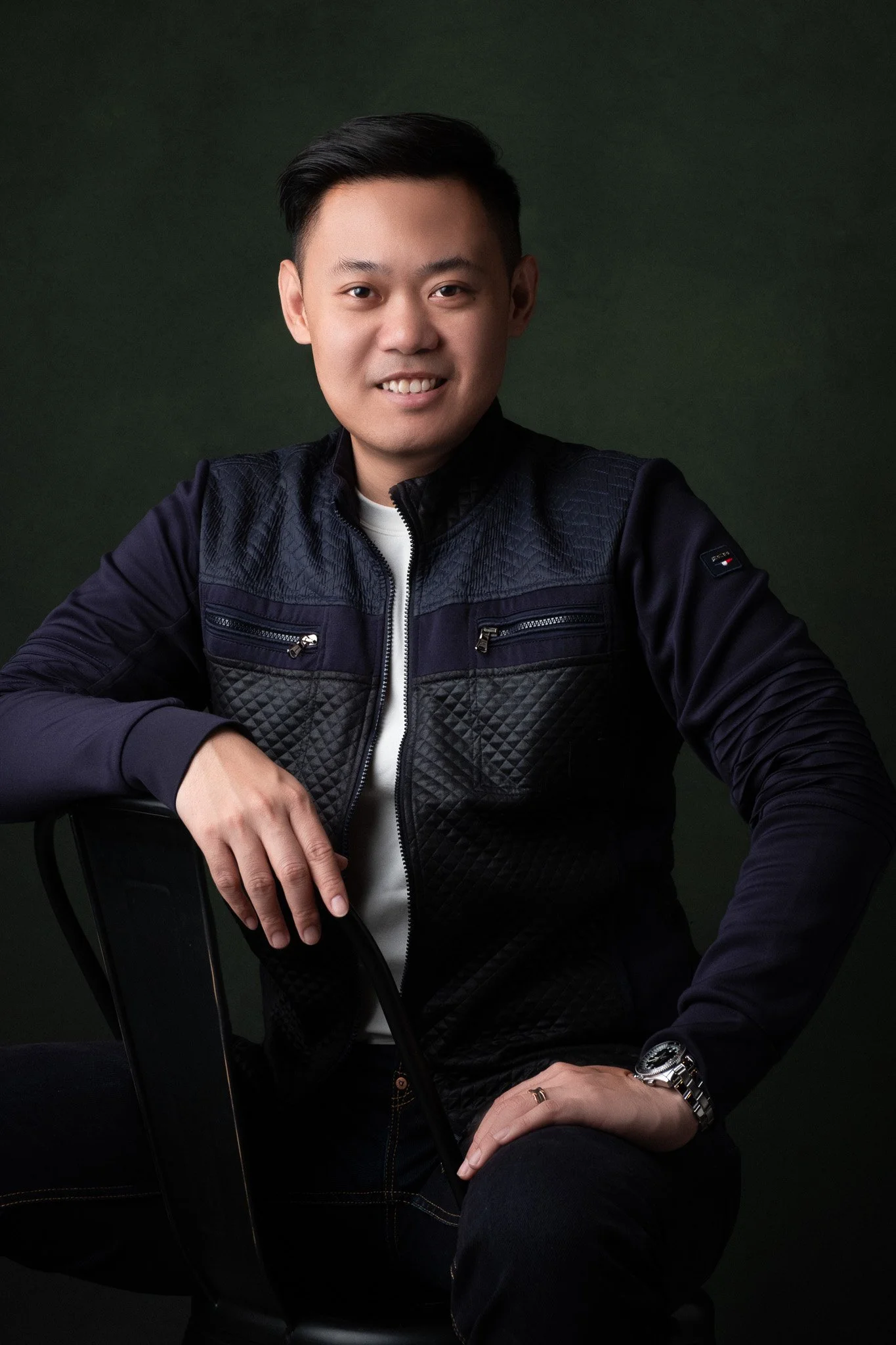 Young man with short black hair smiling, seated wearing a black quilted jacket, white shirt, silver watch, and a ring, against a dark green background.
