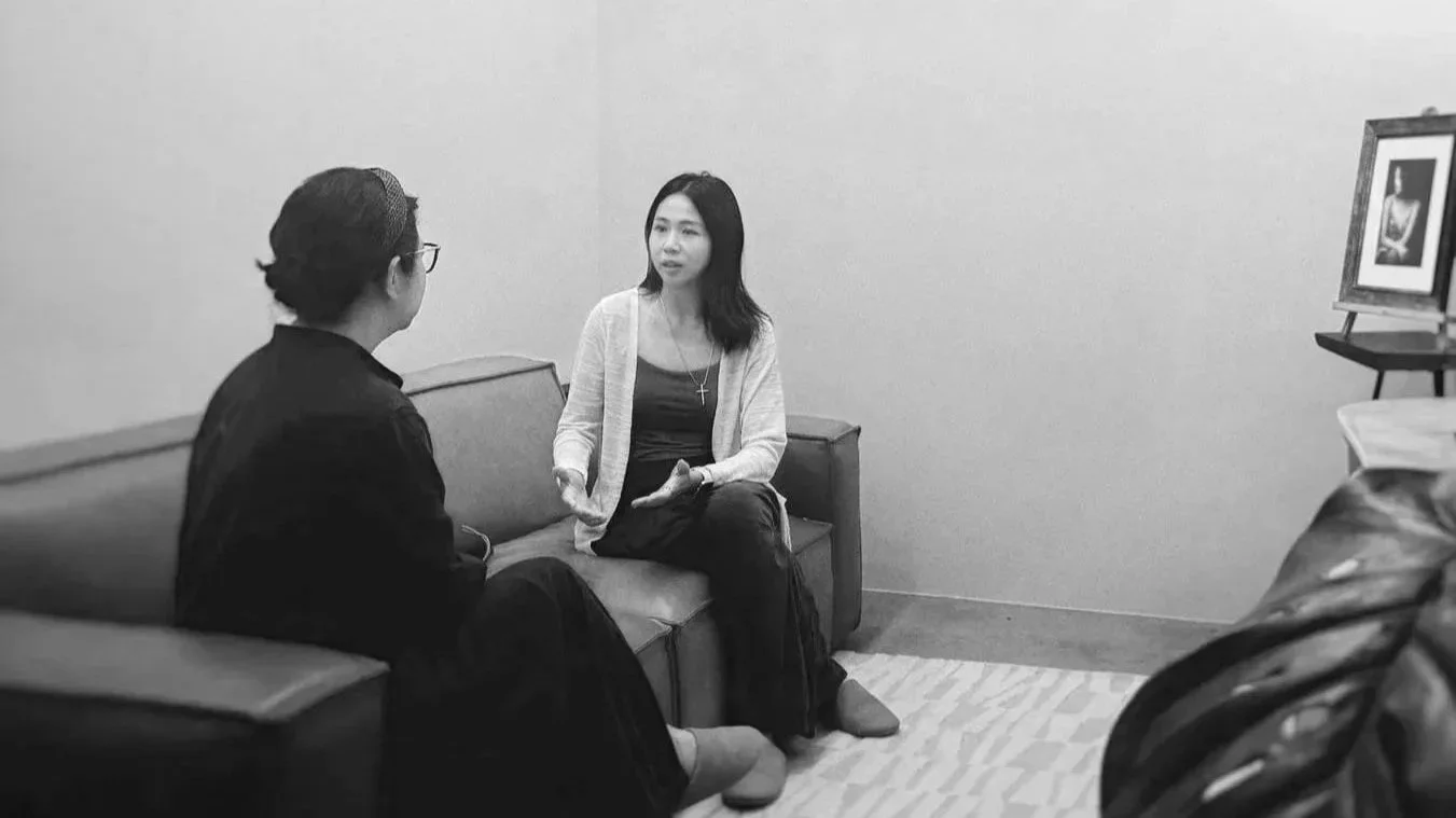 Two women having a conversation in an office setting, seated on couches, with a framed picture on an easel in the background.