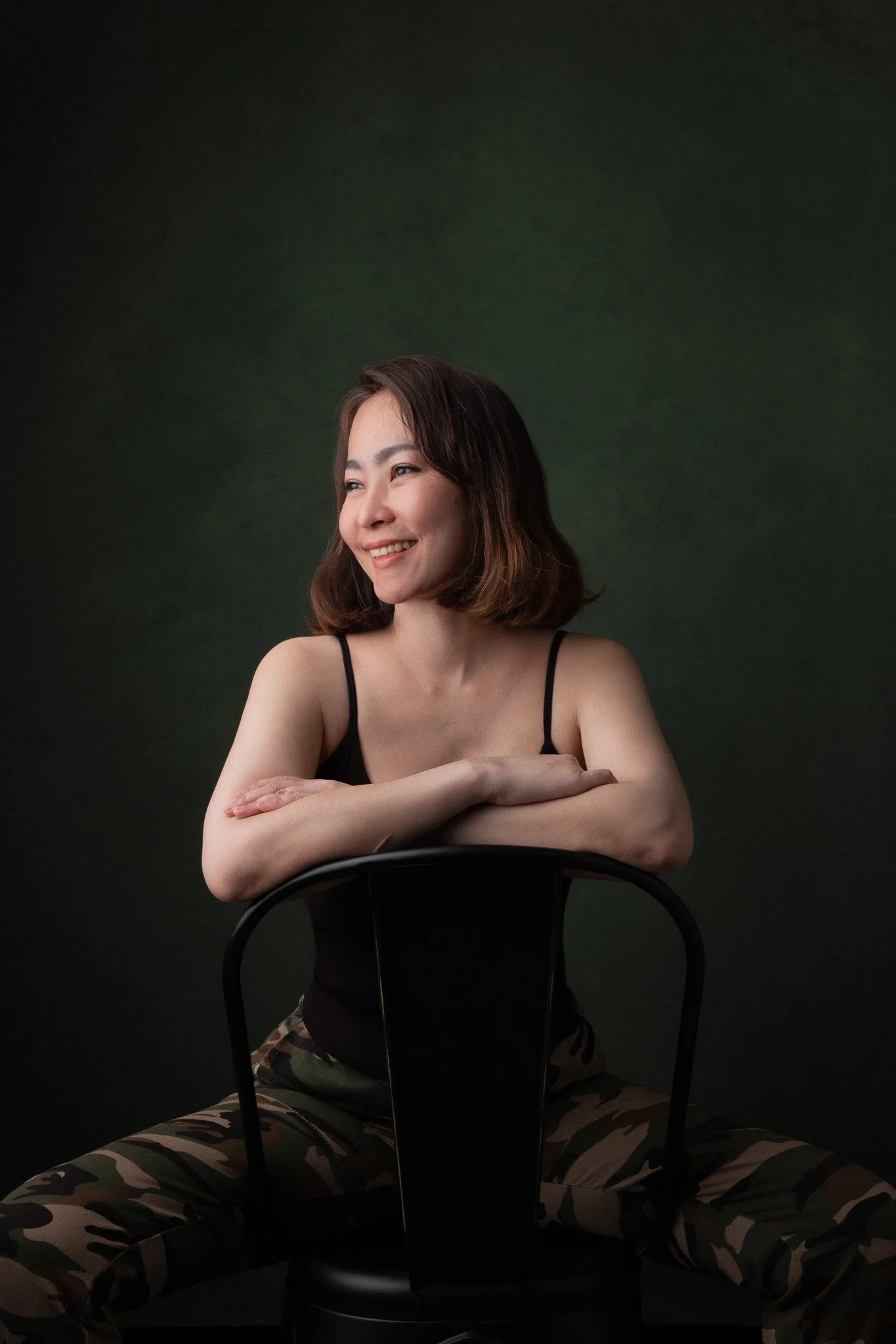 A smiling woman with shoulder-length brown hair sitting cross-legged on the floor, wearing camouflage pants and a black tank top, leaning on a black chair in front of a dark green background.