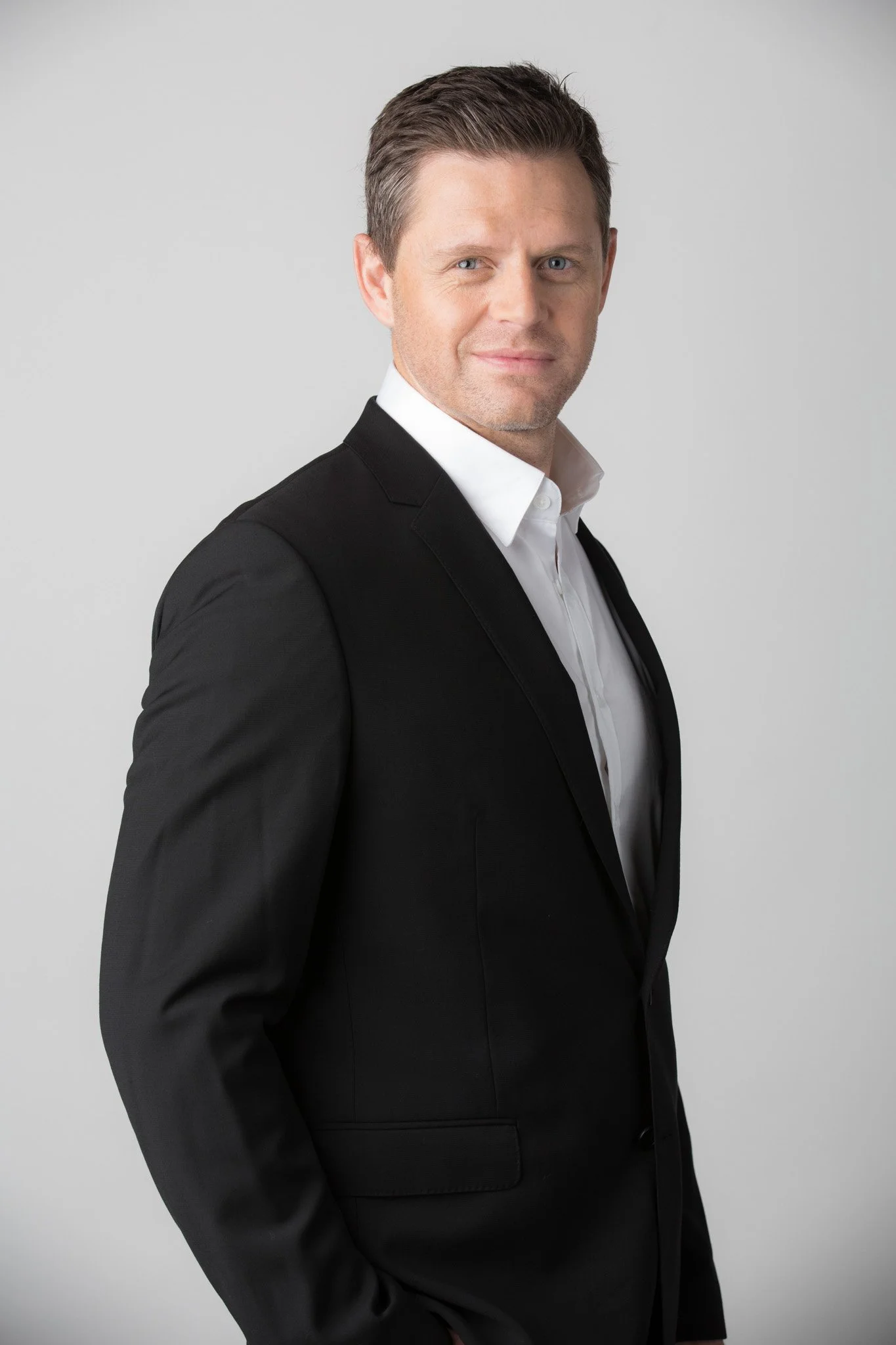 Executive portrait of a man in a black suit with a white shirt stands against a light gray background, winking with a slight smile.