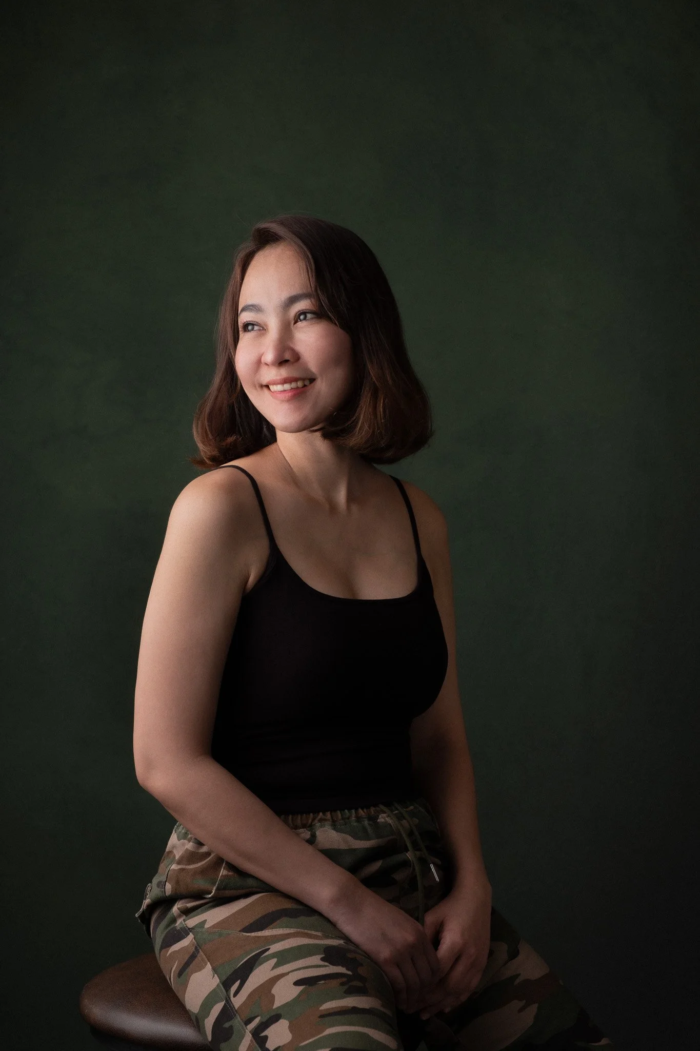 A woman with shoulder-length dark hair, wearing a black spaghetti strap top, sitting on a stool against a dark green backdrop, smiling and looking to her right.