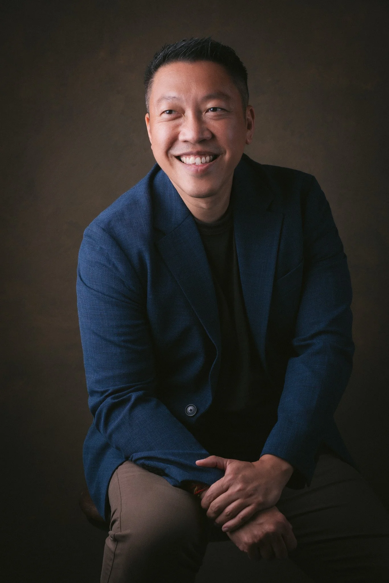 A smiling man with short, dark hair wearing a dark blue blazer and black shirt, sitting against a dark background.