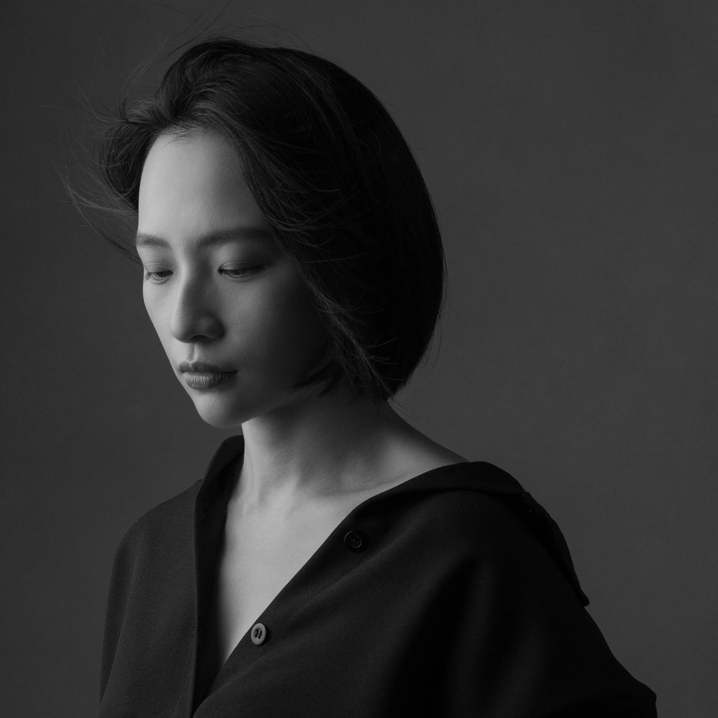 Close-up grayscale photo of a woman's face and upper body, with closed eyes, short dark hair, wearing a dark blazer with a button, near a plain dark background.