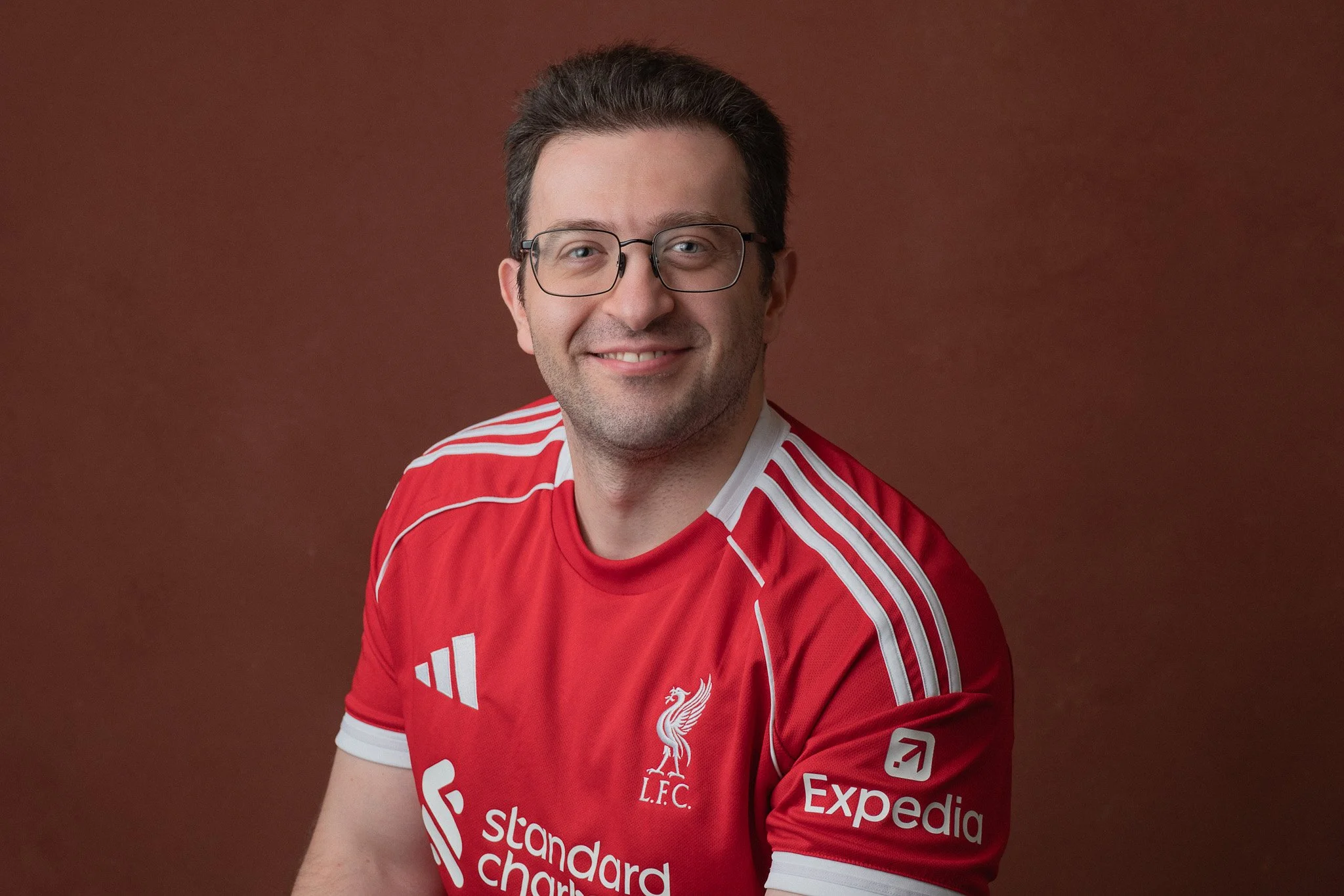 A man wearing glasses and a red Liverpool football jersey smiling at the camera against a brown background.