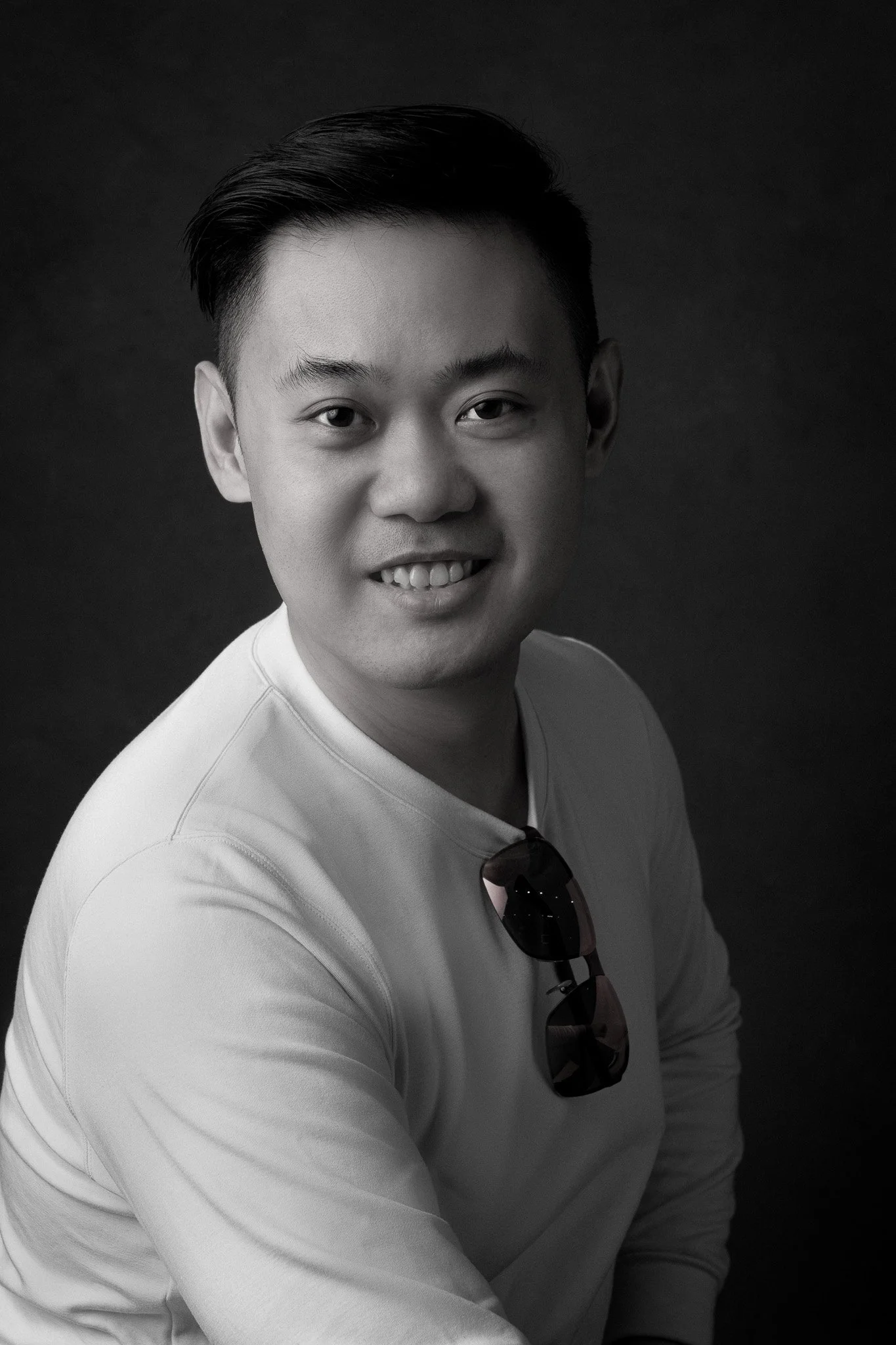 Portrait of a young man with short dark hair, smiling slightly, wearing a light-colored long sleeve shirt, with sunglasses hanging from his shirt collar against a dark background.
