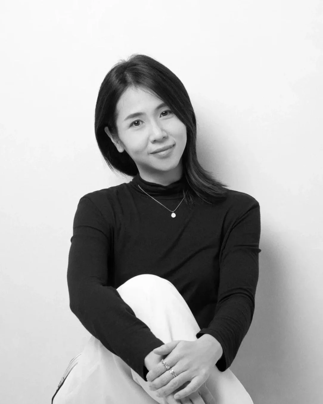 A woman with shoulder-length dark hair, wearing a black long-sleeve top and a necklace, sitting against a plain white wall, smiling softly and looking at the camera.