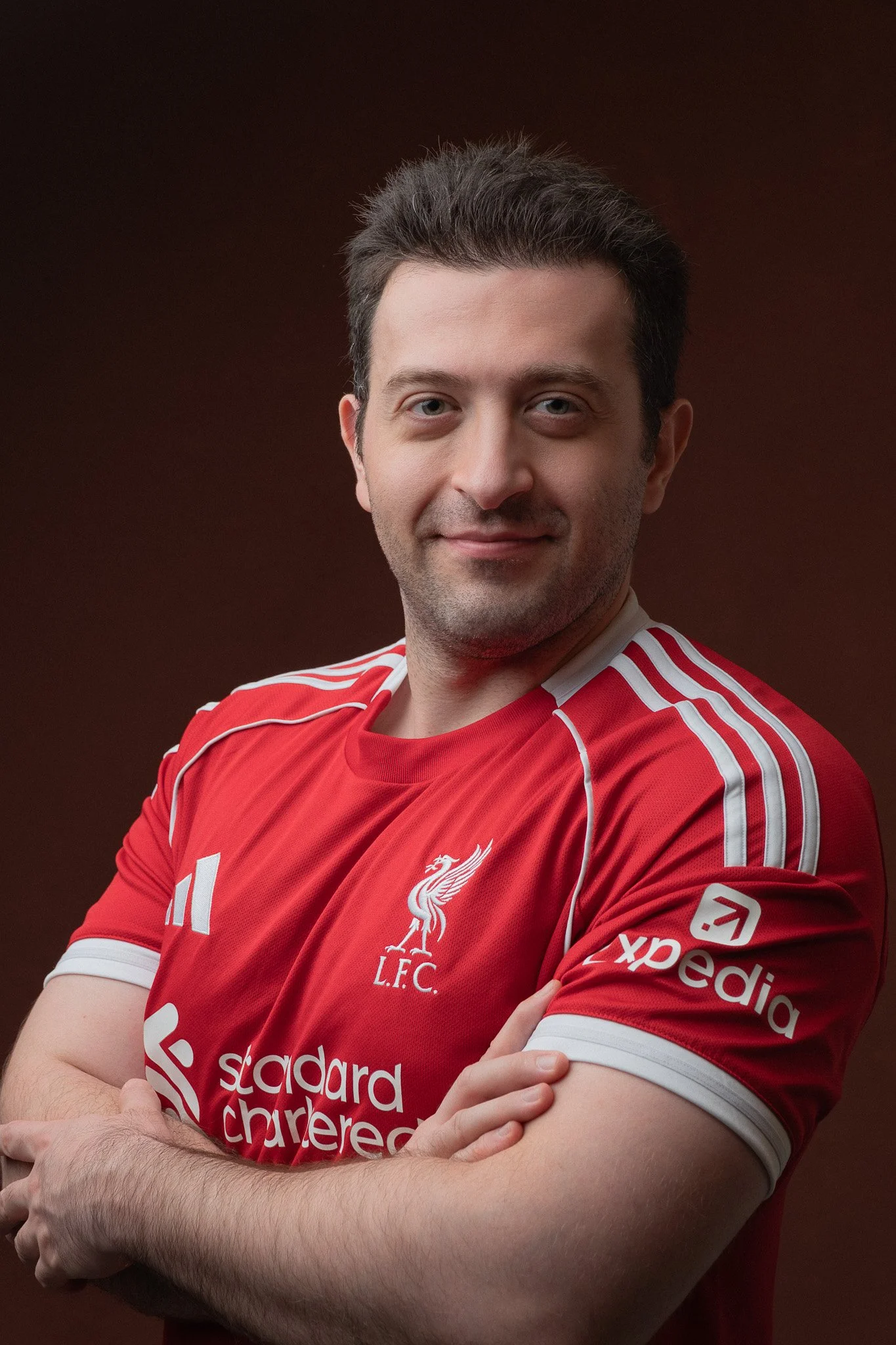 Close-up of a man in a red Liverpool Football Club jersey with arms crossed, smiling slightly, against a dark background.