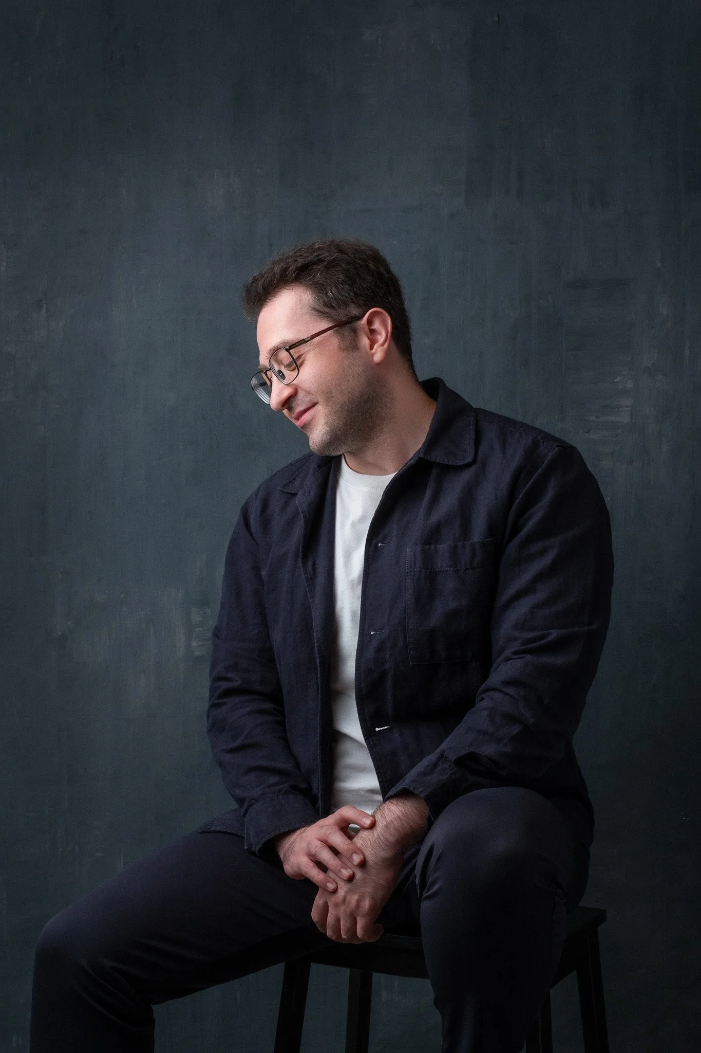 A young man with dark hair, glasses, and light skin sitting on a black stool against a dark wall, smiling with eyes closed, dressed in a white T-shirt and a dark jacket.