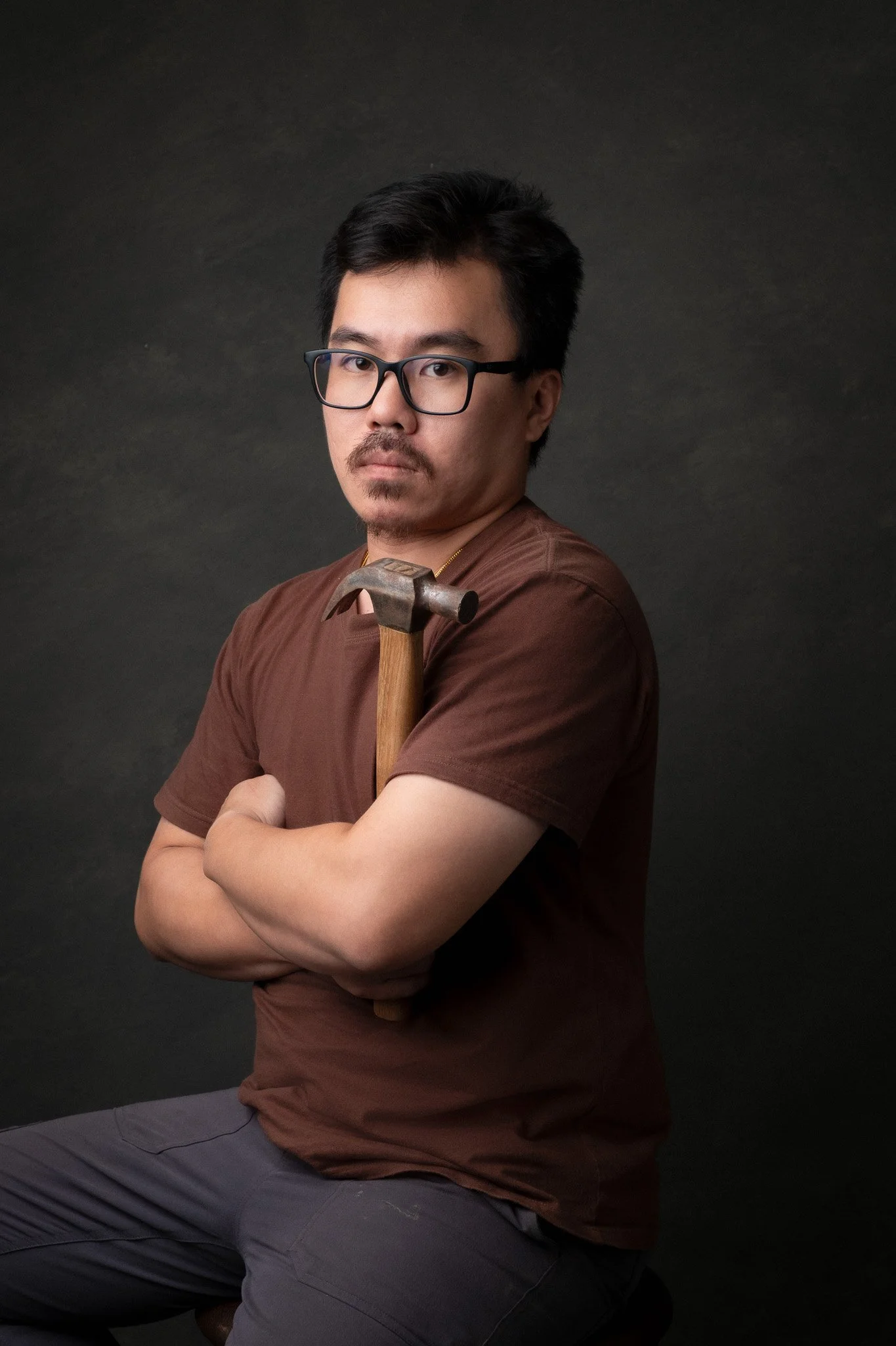 A man with black hair, glasses, and a mustache, sitting with arms crossed, holding a hammer on his shoulder, wearing a brown t-shirt and gray pants, against a dark background.
