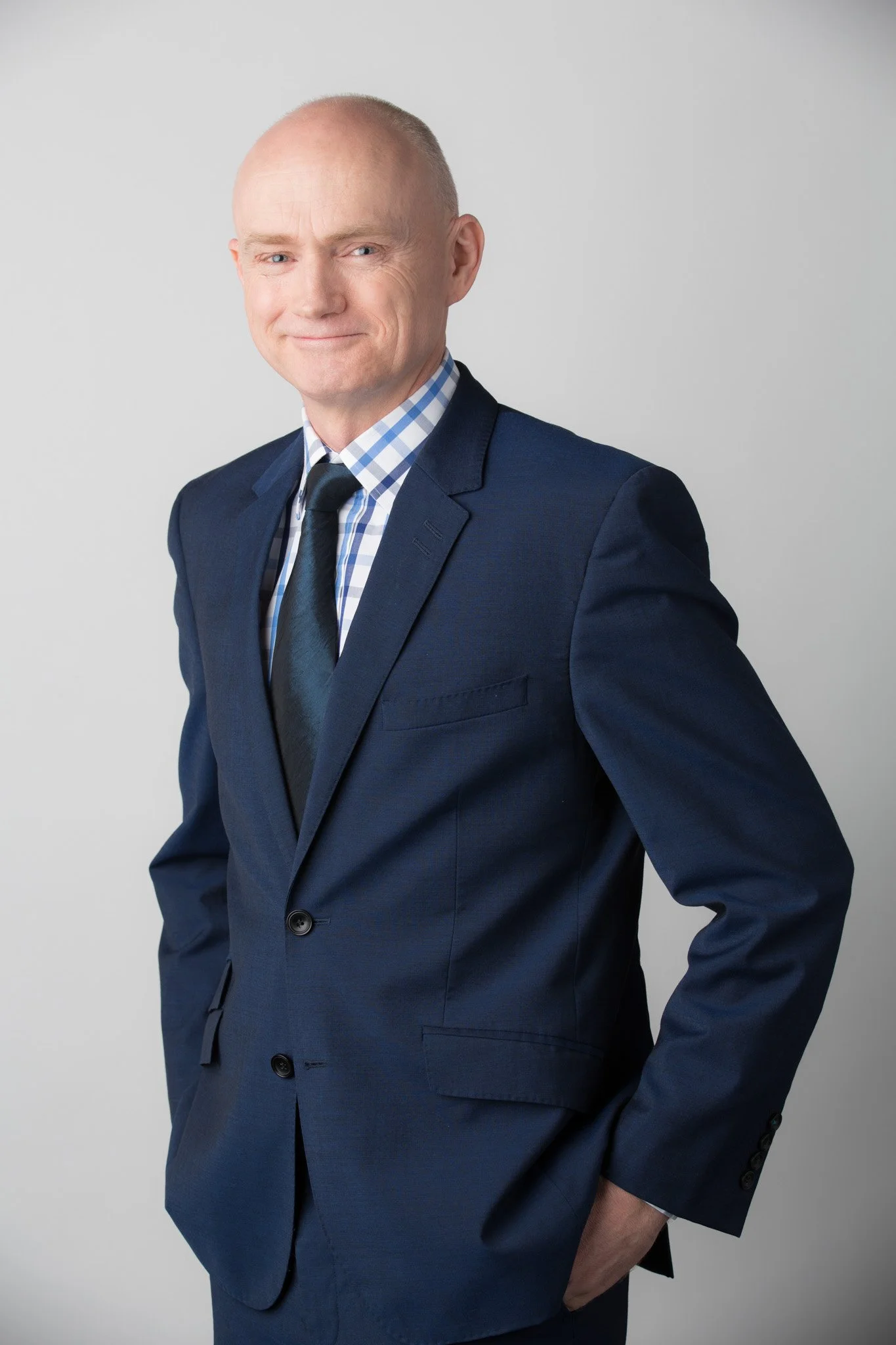 Corporate headshot of a middle-aged man in a dark blue suit with a checkered shirt and a dark tie, smiling slightly with one arm bent and his hand in his pocket, standing against a plain gray background.
