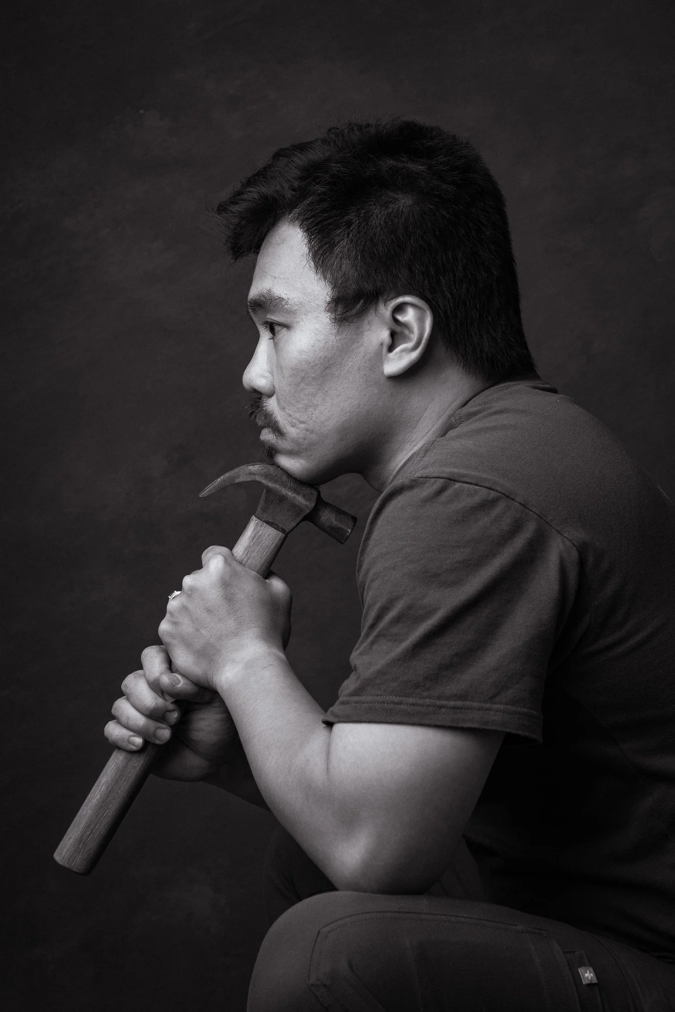 A young man with dark hair and a mustache holding a hammer with both hands against a dark background.
