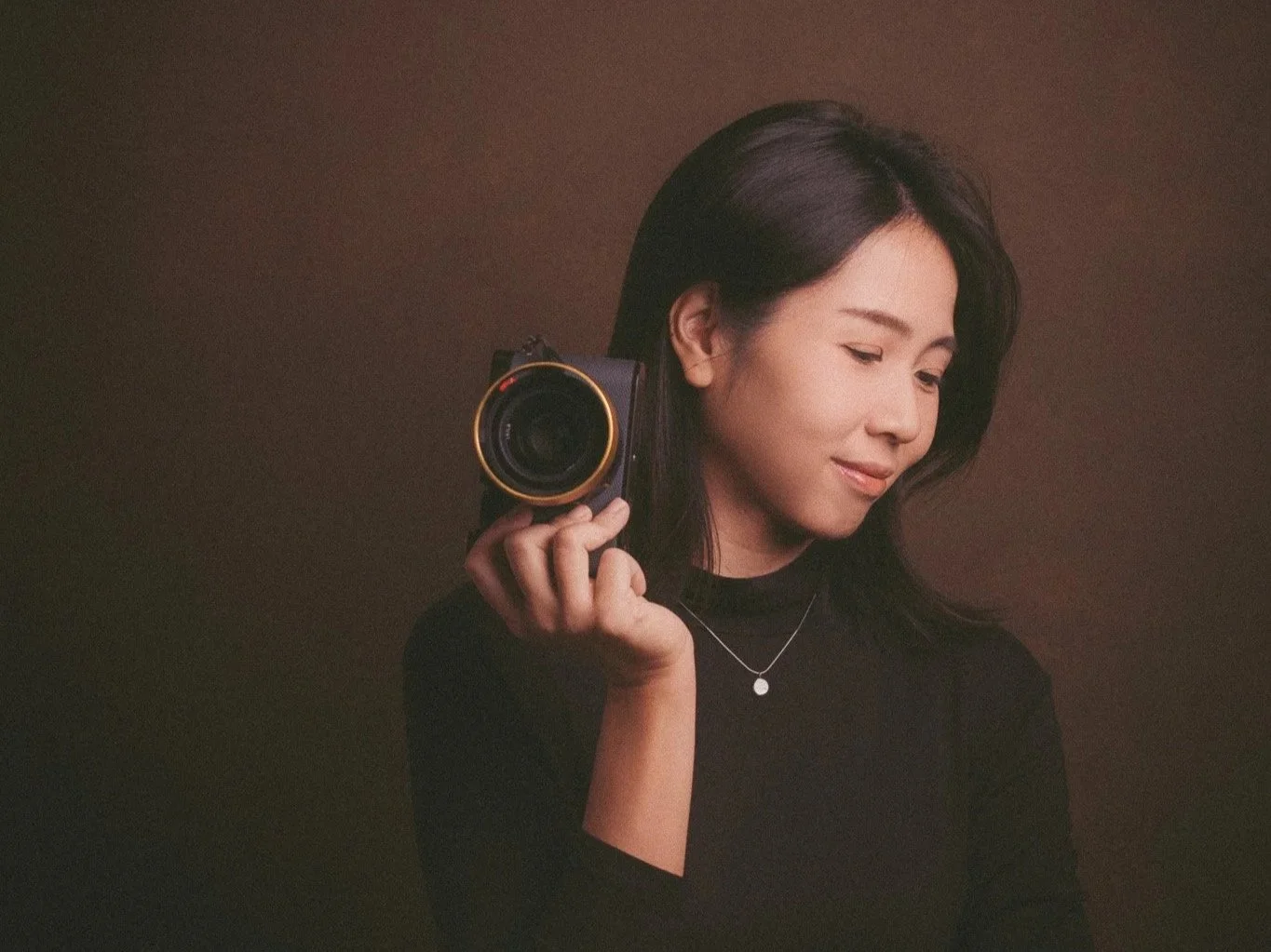 A woman with dark hair holding a camera resting on her shoulder, wearing a black top and a necklace, against a brown background.