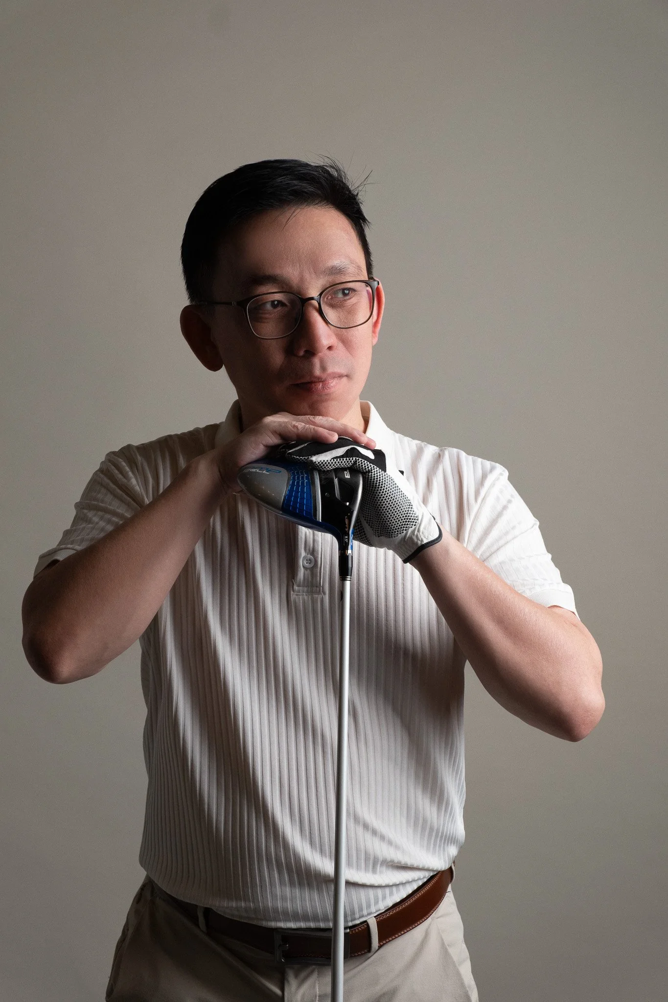 A man with glasses wearing a white dress shirt, holding a blue and black golf club, looking to the side, against a neutral background.
