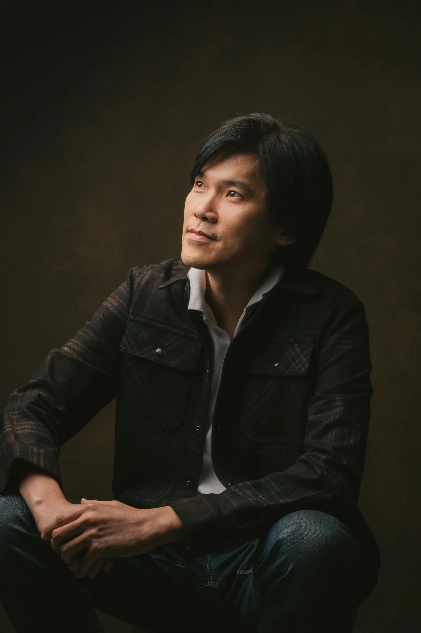 A man with black hair sitting against a brown background, looking to his right with a thoughtful expression, wearing a dark checked jacket and a white shirt.