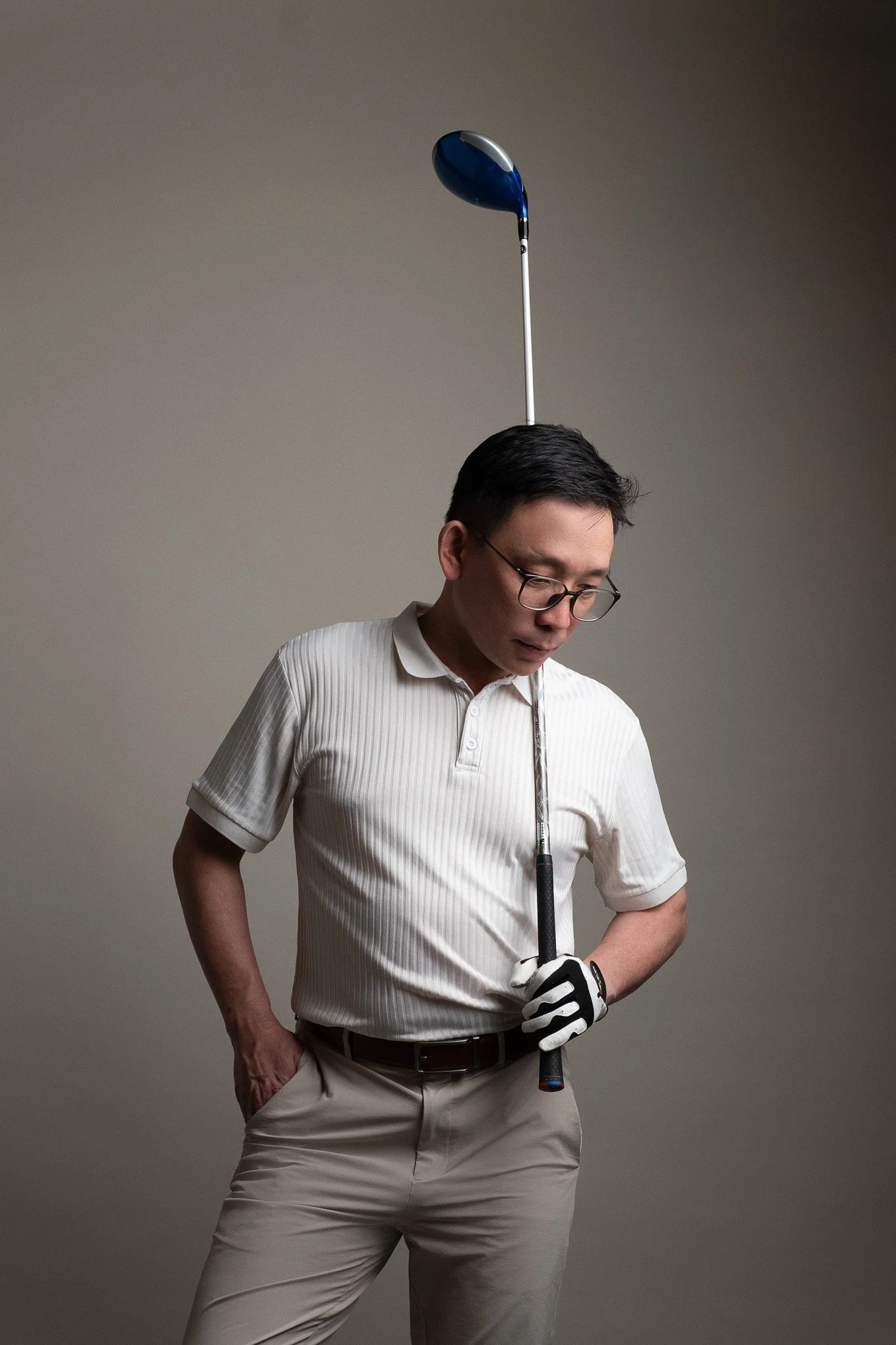 A man wearing glasses and a white polo shirt holding a golf club with a golf glove on his left hand, with a golf ball in his pocket, against a plain background.
