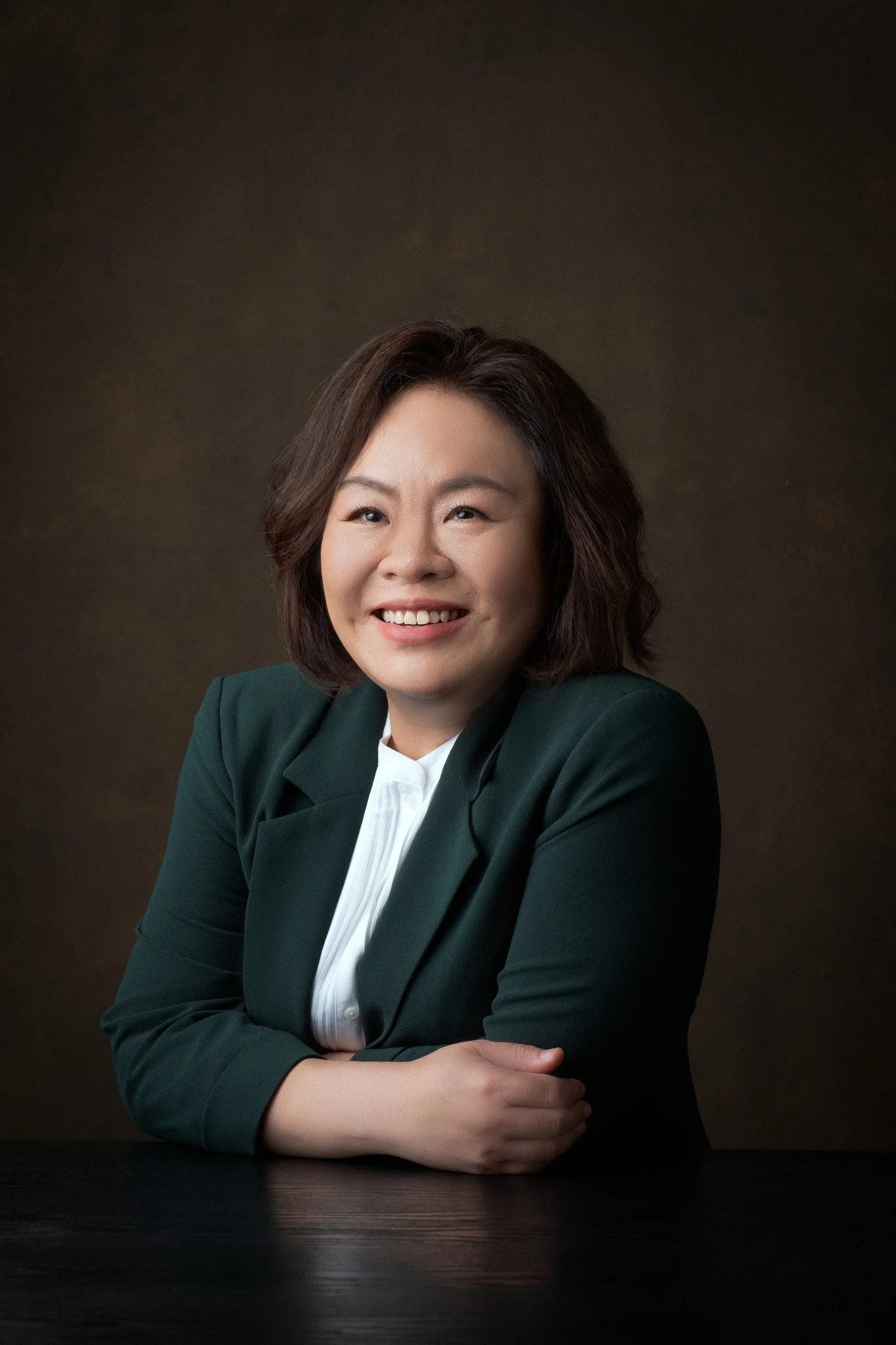 Headshot of a smiling Asian woman in a dark green blazer and white shirt, sitting at a black table against a dark background.