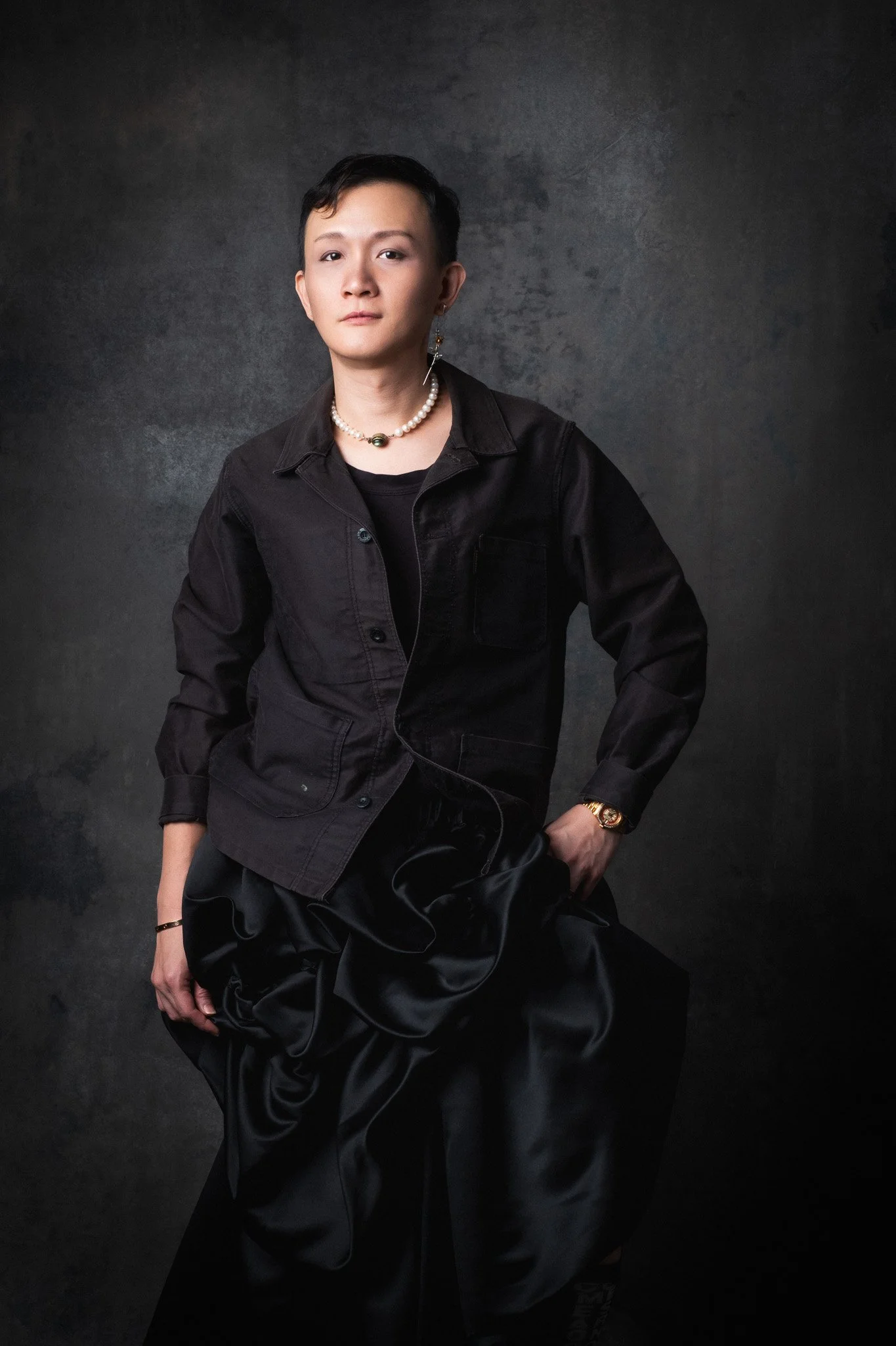A person with short dark hair wearing a black jacket, black satin-like pants, and jewelry including a pearl necklace and earrings, posing against a dark textured background.
