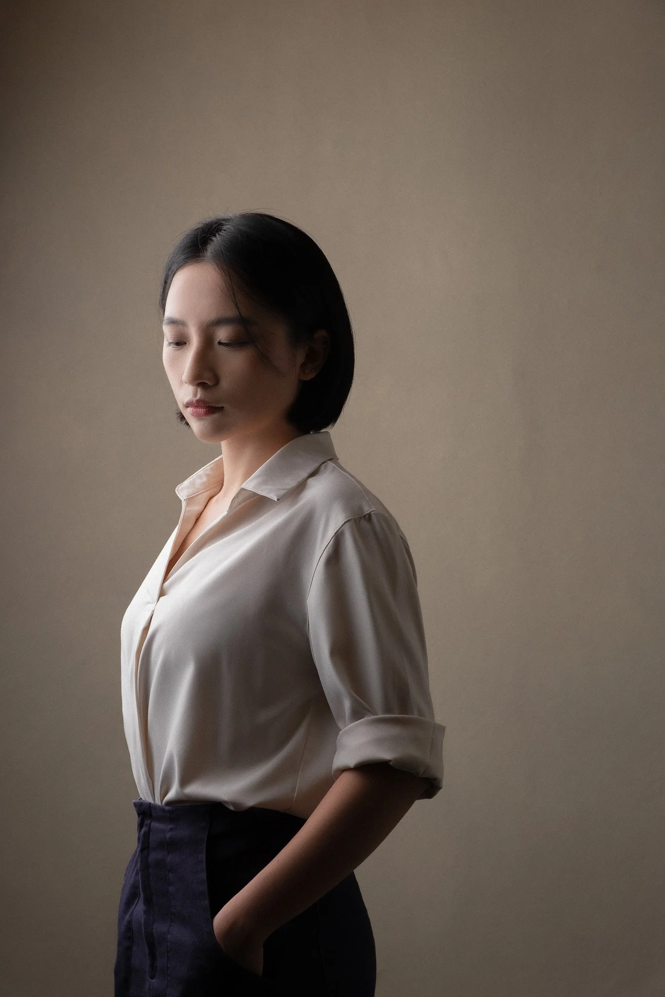 A woman with short black hair wearing a beige blouse and dark pants, standing in front of a plain, neutral background.