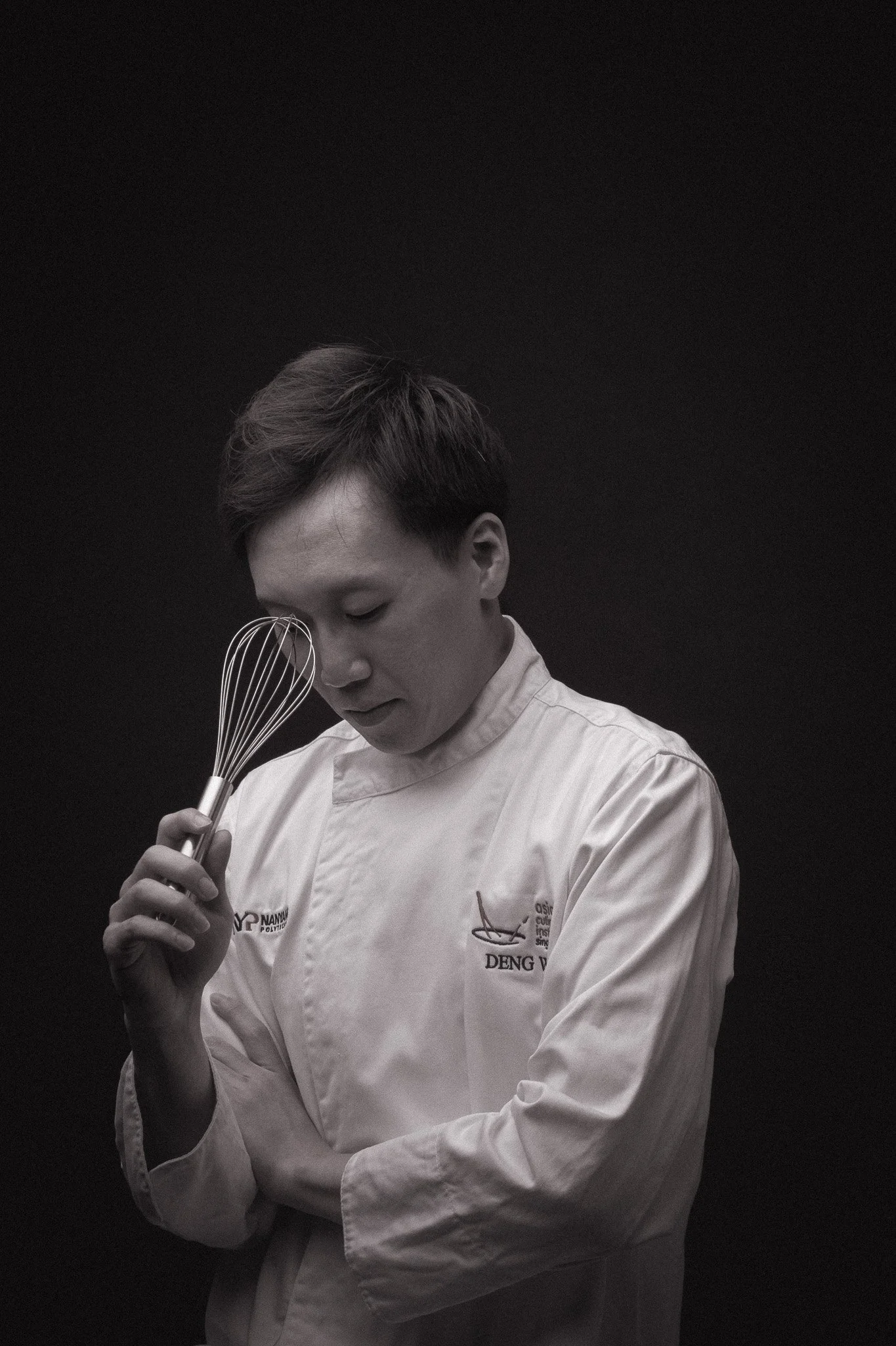 Black and white photo of a person in a chef's uniform holding a whisk, looking down, against a black background.