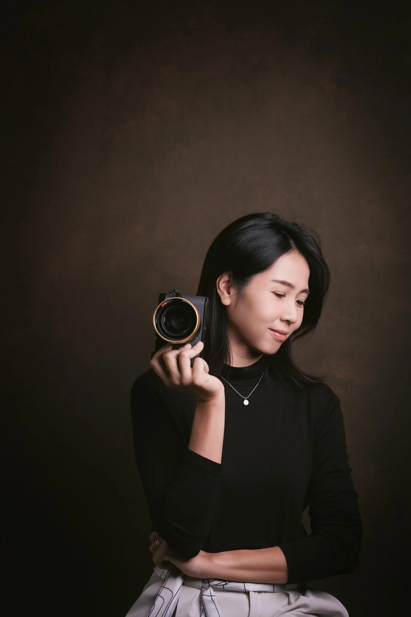 A woman with long black hair holding a black camera near her face, wearing a black top and jewelry, with a neutral expression against a dark background.
