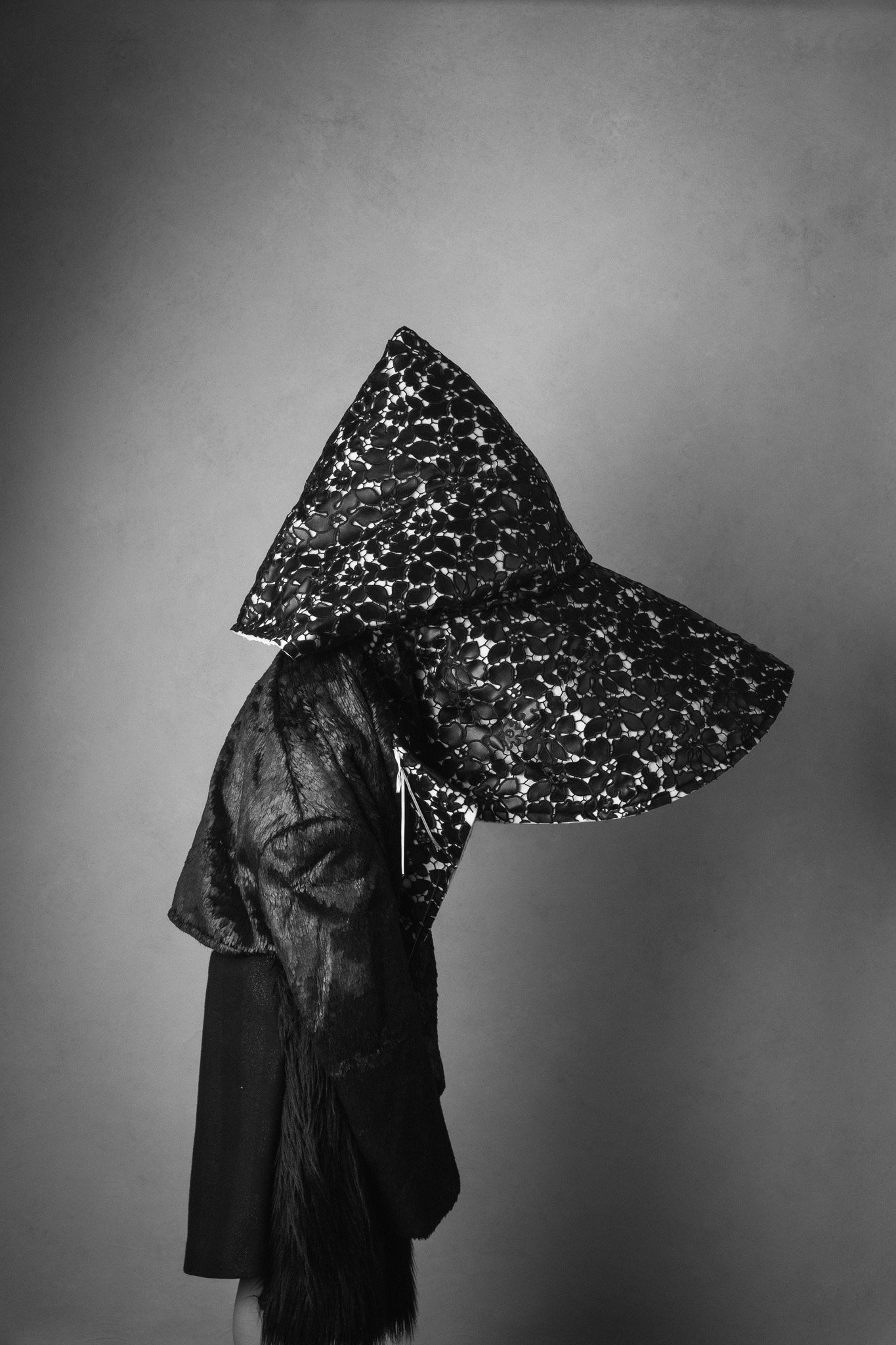 A person standing sideways wearing a large, decorative, open-cut hat with a pattern resembling linked flowers, and a loose, textured jacket with a fur or leather section on one sleeve.