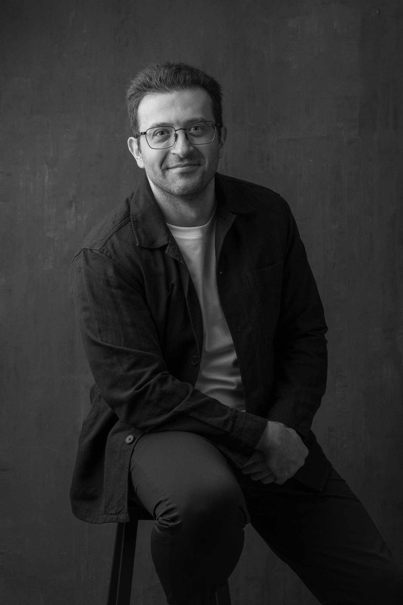 Black and white portrait of a man wearing glasses, a white T-shirt, and a dark jacket, sitting on a chair against a plain background.