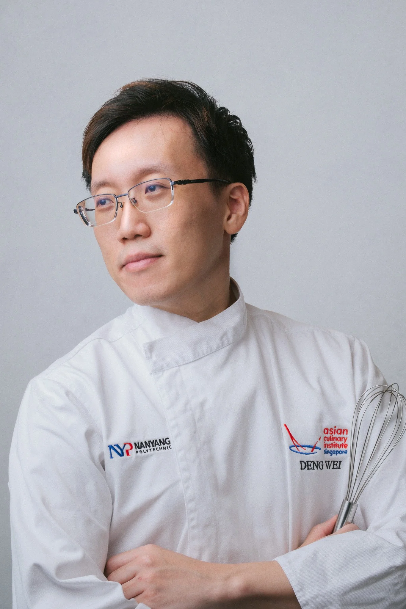 A young man with glasses wearing a white chef's coat with logos from Nanyang Polytechnic and the Asian Culinary Institute Singapore, holding a wire whisk.