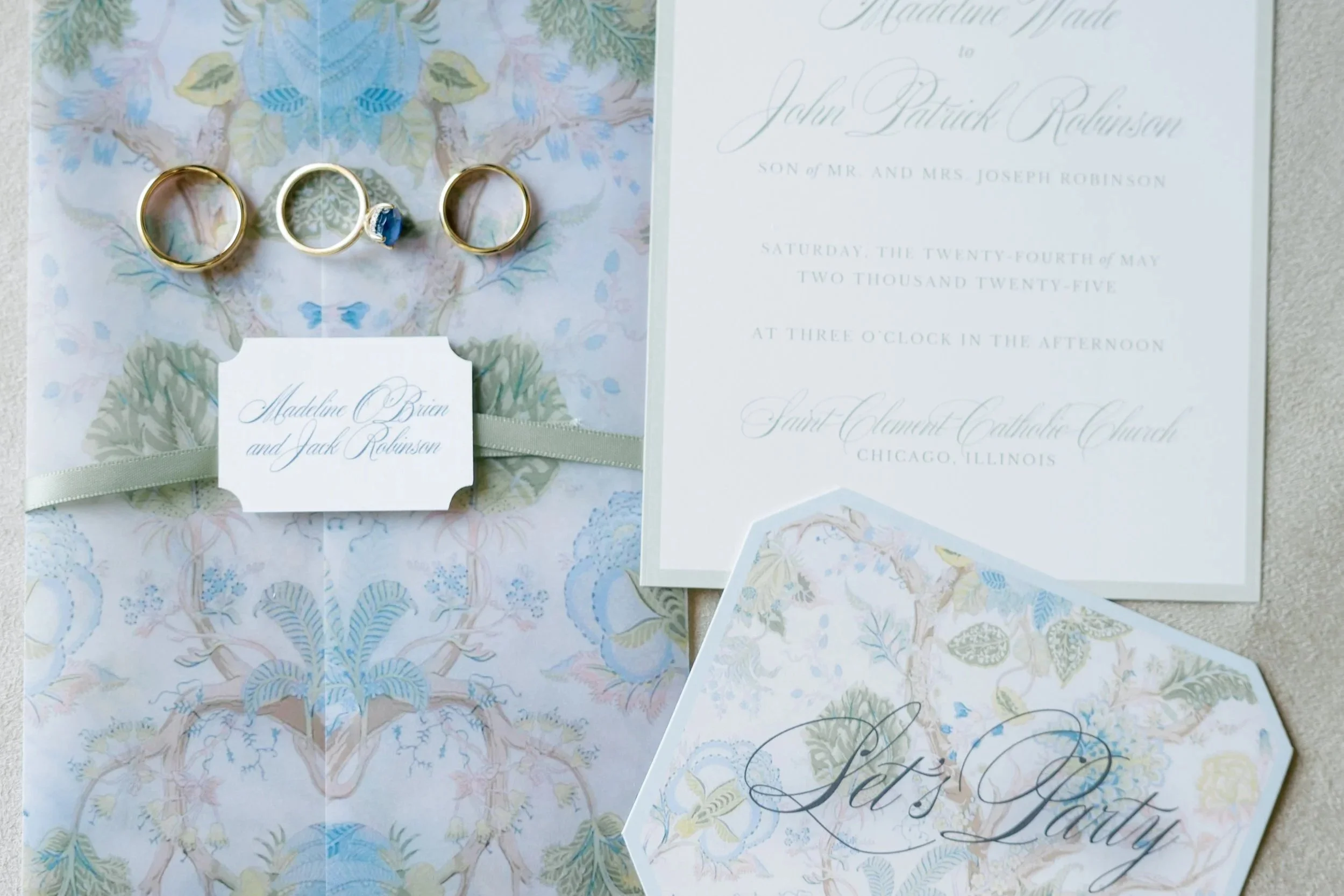 Wedding invitation suite with three gold rings, a blue gemstone ring, and a label with names Madeline Brain and Jack Robison, all decorated with pale blue, cream, and pastel-colored floral patterns.