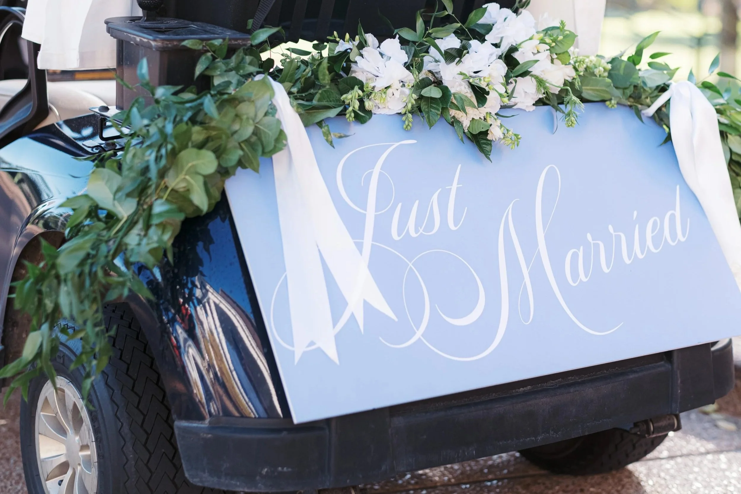 A decorated golf cart with a light blue sign on the side reading 'Just Married,' adorned with white flowers and green foliage.