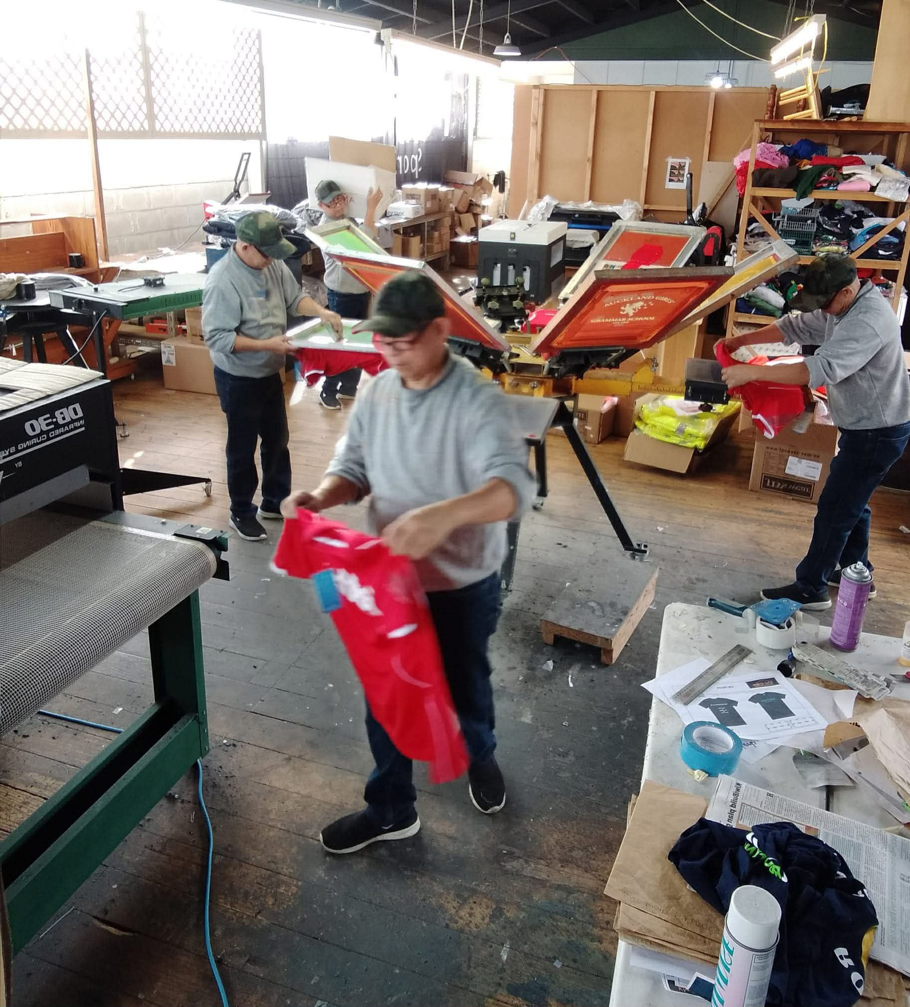 Workers in a screen printing workshop preparing and printing red T-shirts, with printing machines, supplies, and design plans visible.