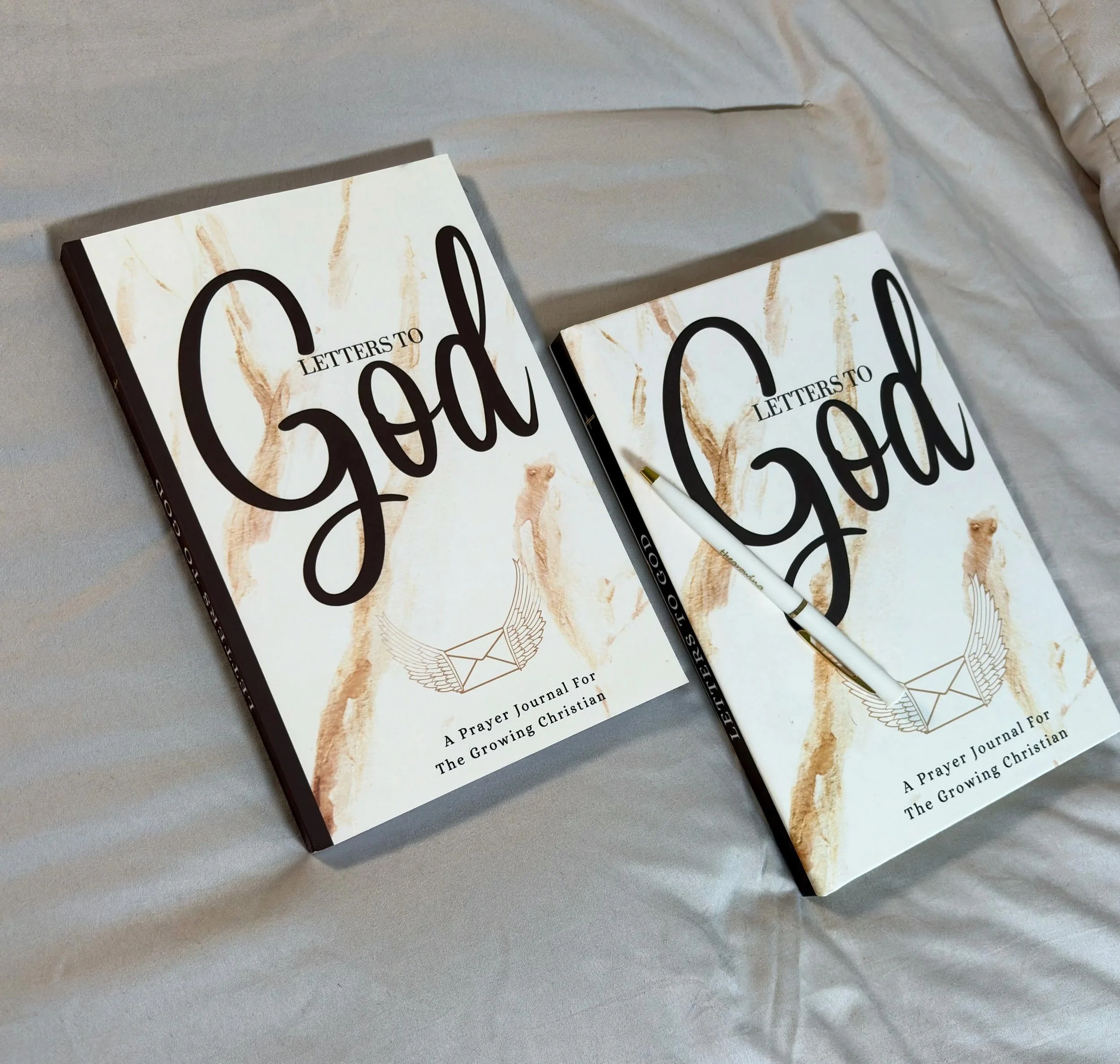 Two books titled "Letters to God" with a white pen resting on the open book, placed on a light-colored fabric surface.