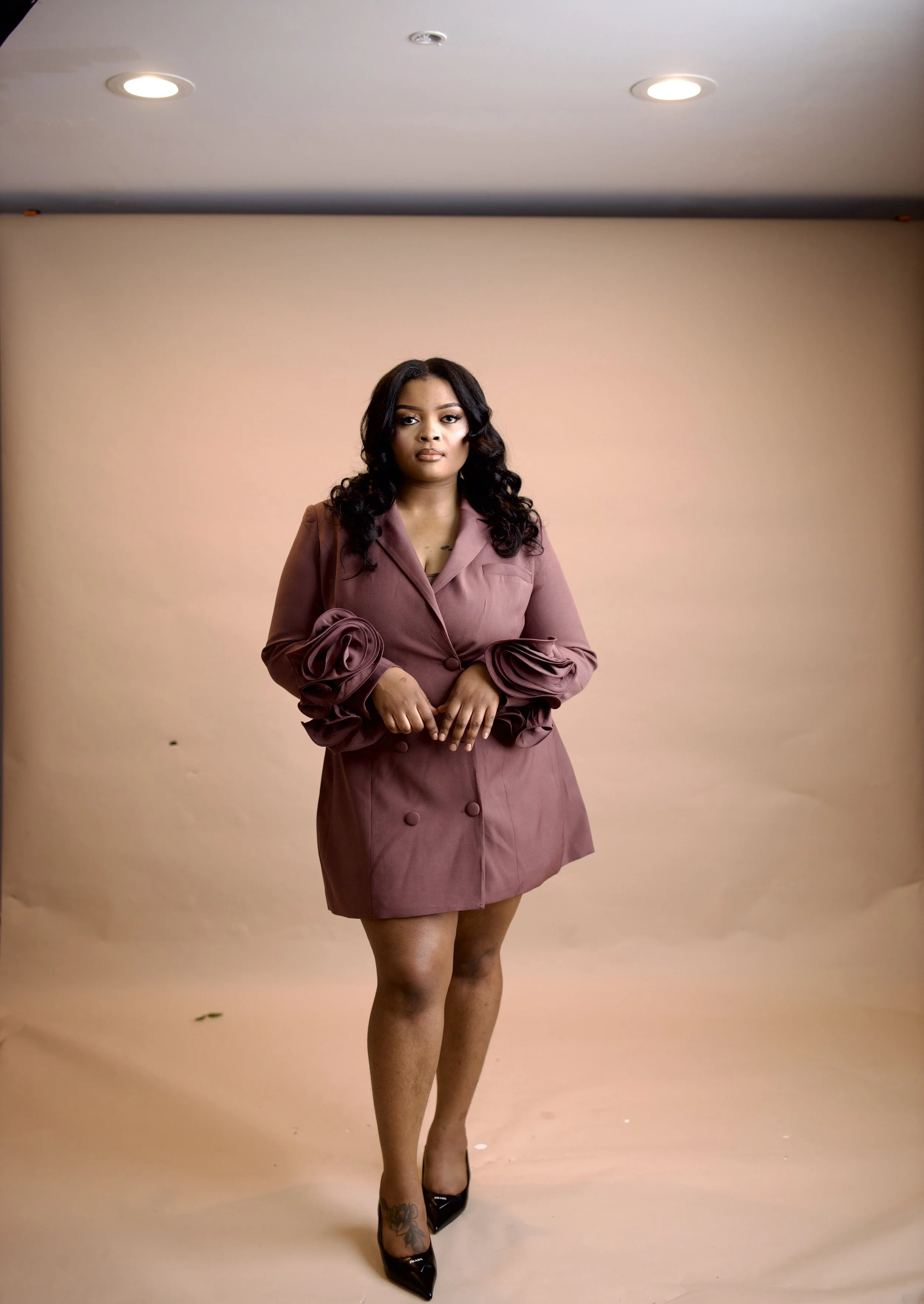 A woman with long curly black hair wearing a mauve blazer with oversized ribbon-like sleeves and matching skirt, black heels, standing in front of a beige backdrop in a photo studio.