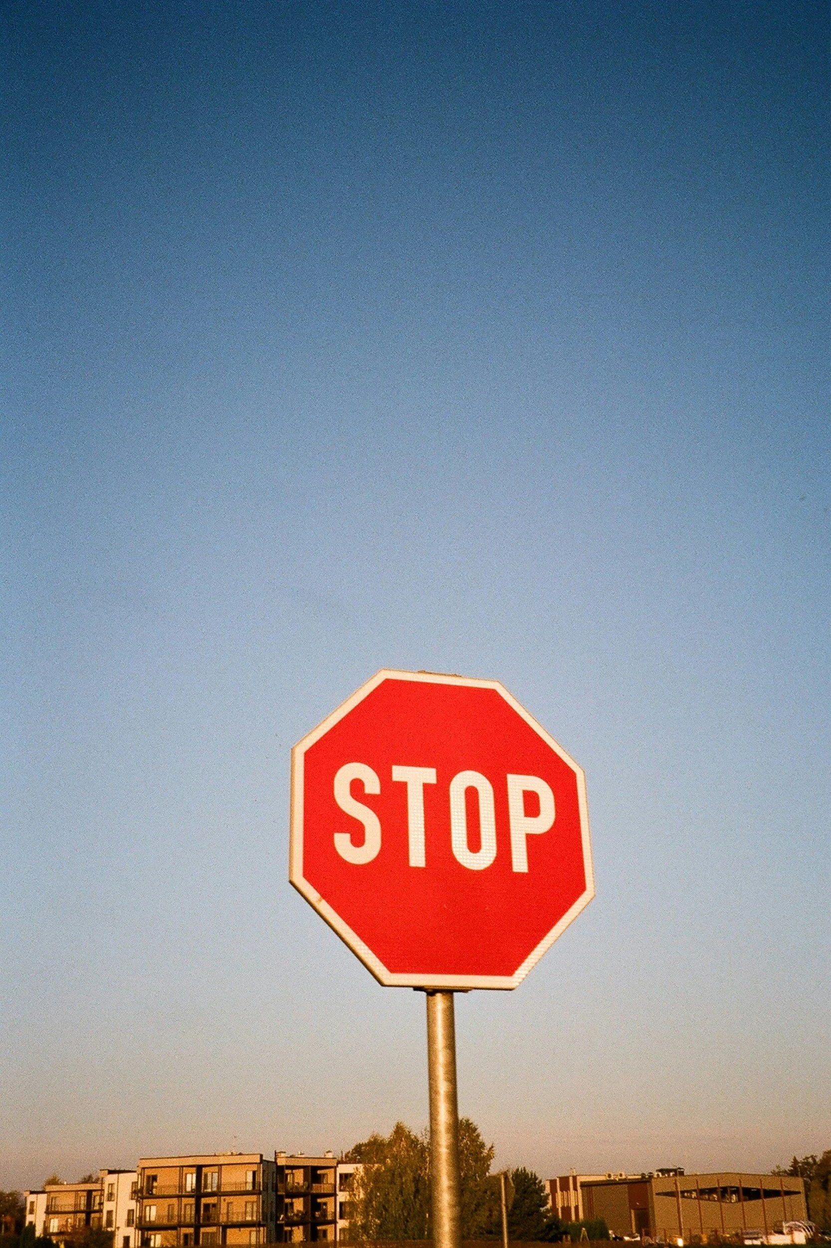 A red stop sign with white letters stands against a clear blue sky, with modern apartment buildings and trees in the background.