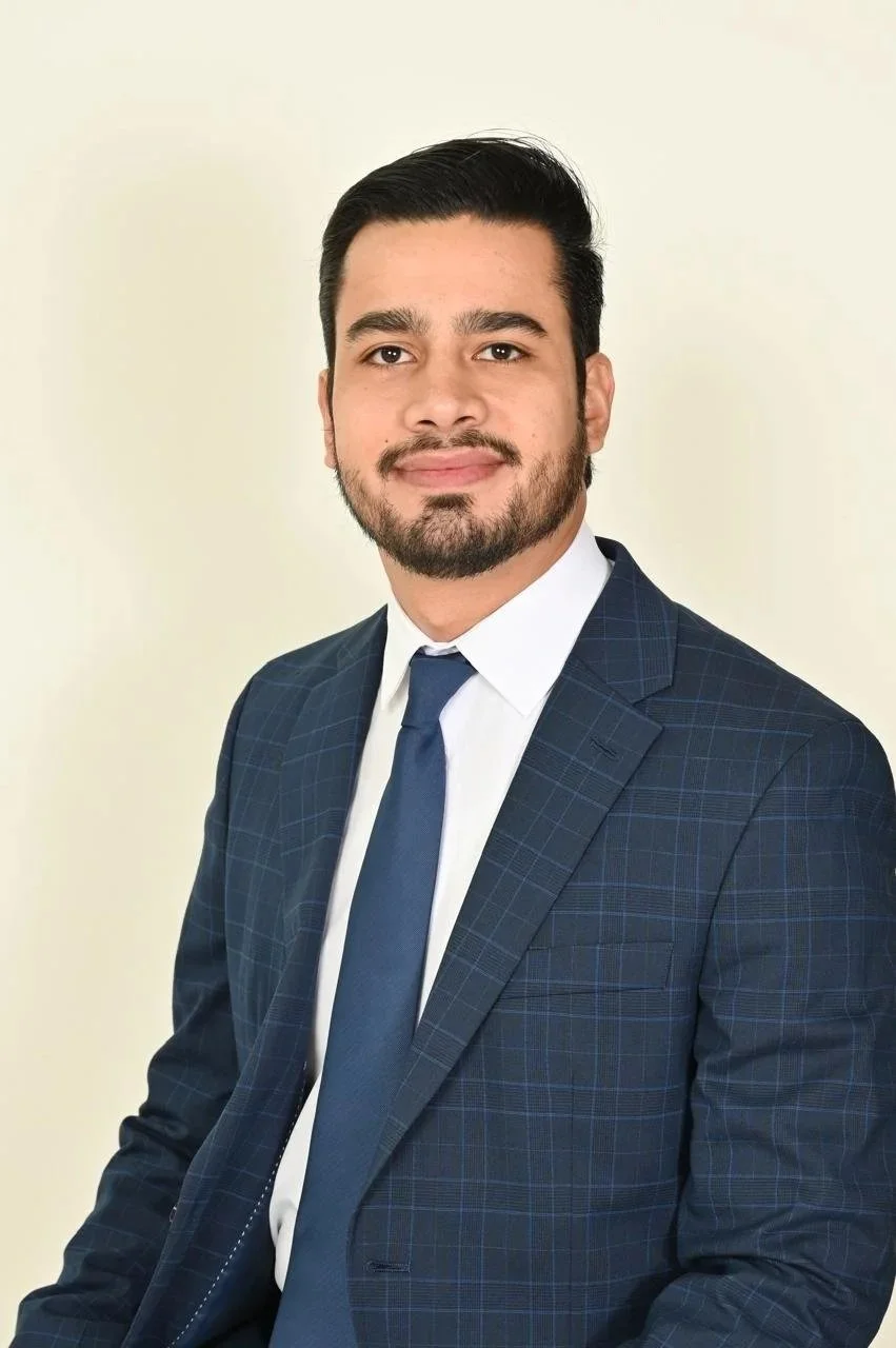 A man in a dark blue checked suit, white shirt, and blue tie, with dark hair and a beard, posing for a professional portrait against a light background.