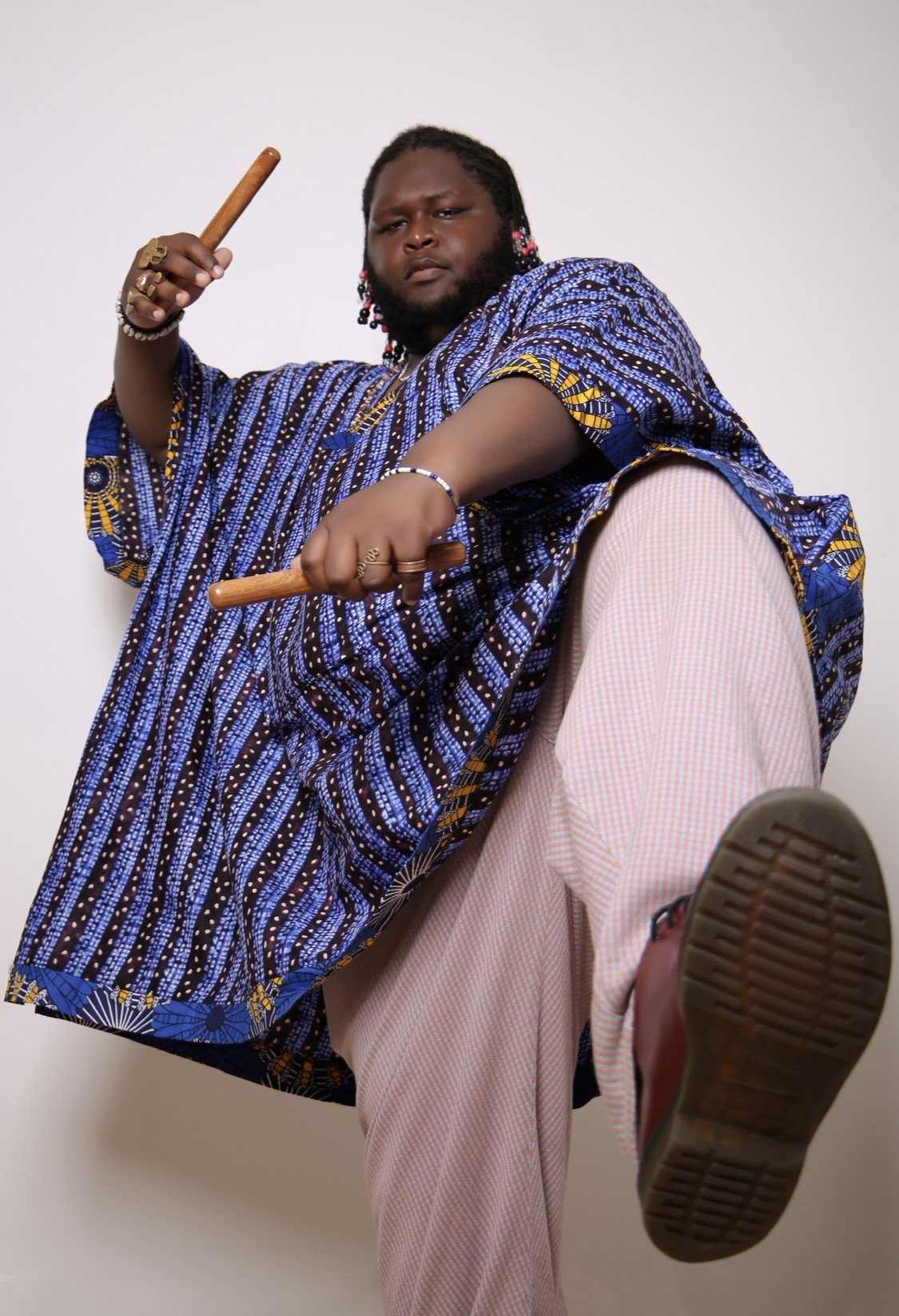 A person wearing traditional African clothing, holding two wooden sticks, and triumphantly kicking a foot forward.