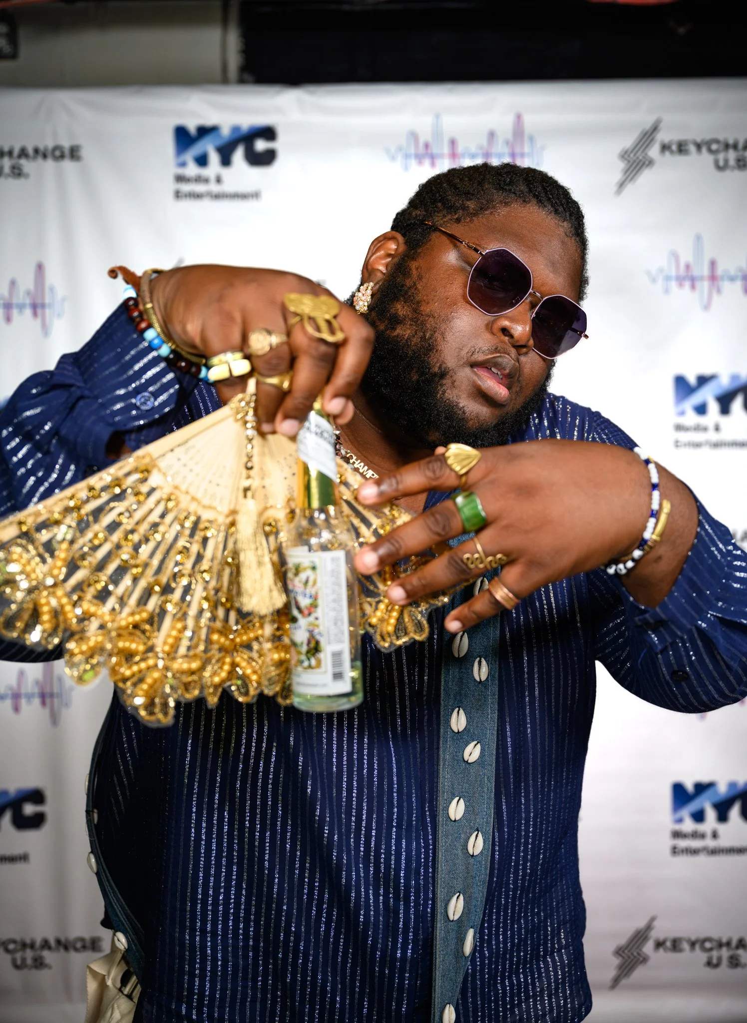 A man wearing dark sunglasses, a blue striped shirt with shell buttons, and numerous gold jewelry pieces, holding a decorated fan and a small bottle, posing in front of a backdrop with logos and sound wave graphics.
