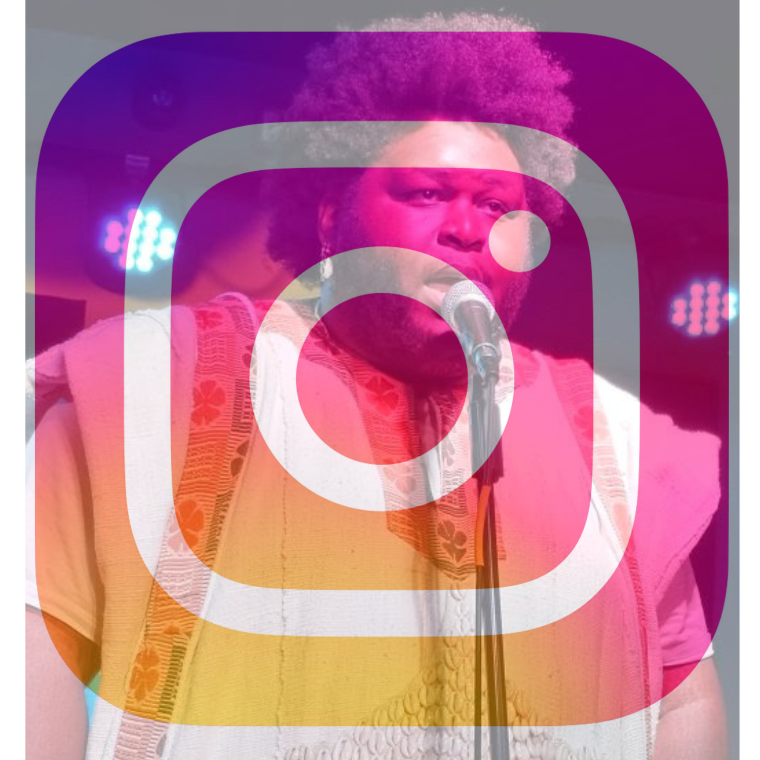 A person with an afro hairstyle and a beard, wearing a beige traditional shirt, speaking into a microphone on stage with colored lights in the background.