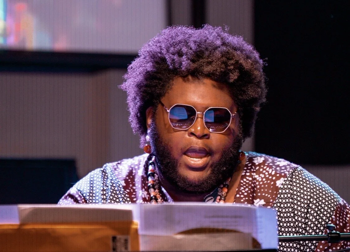 A person with an afro hairstyle wearing sunglasses, earrings, and colorful clothing, sitting in front of a keyboard with a background of bookshelves.