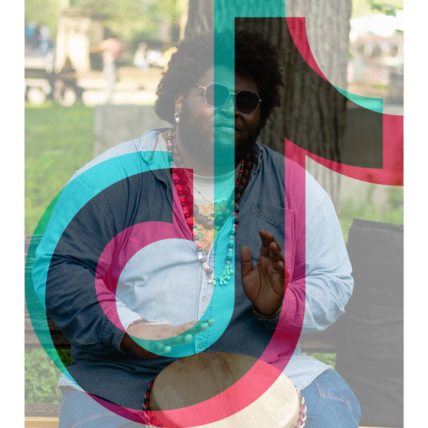 A person sitting on a park bench, playing a drum. They have sunglasses, a denim shirt, colorful necklaces, and are outdoors with trees and blurred buildings in the background.