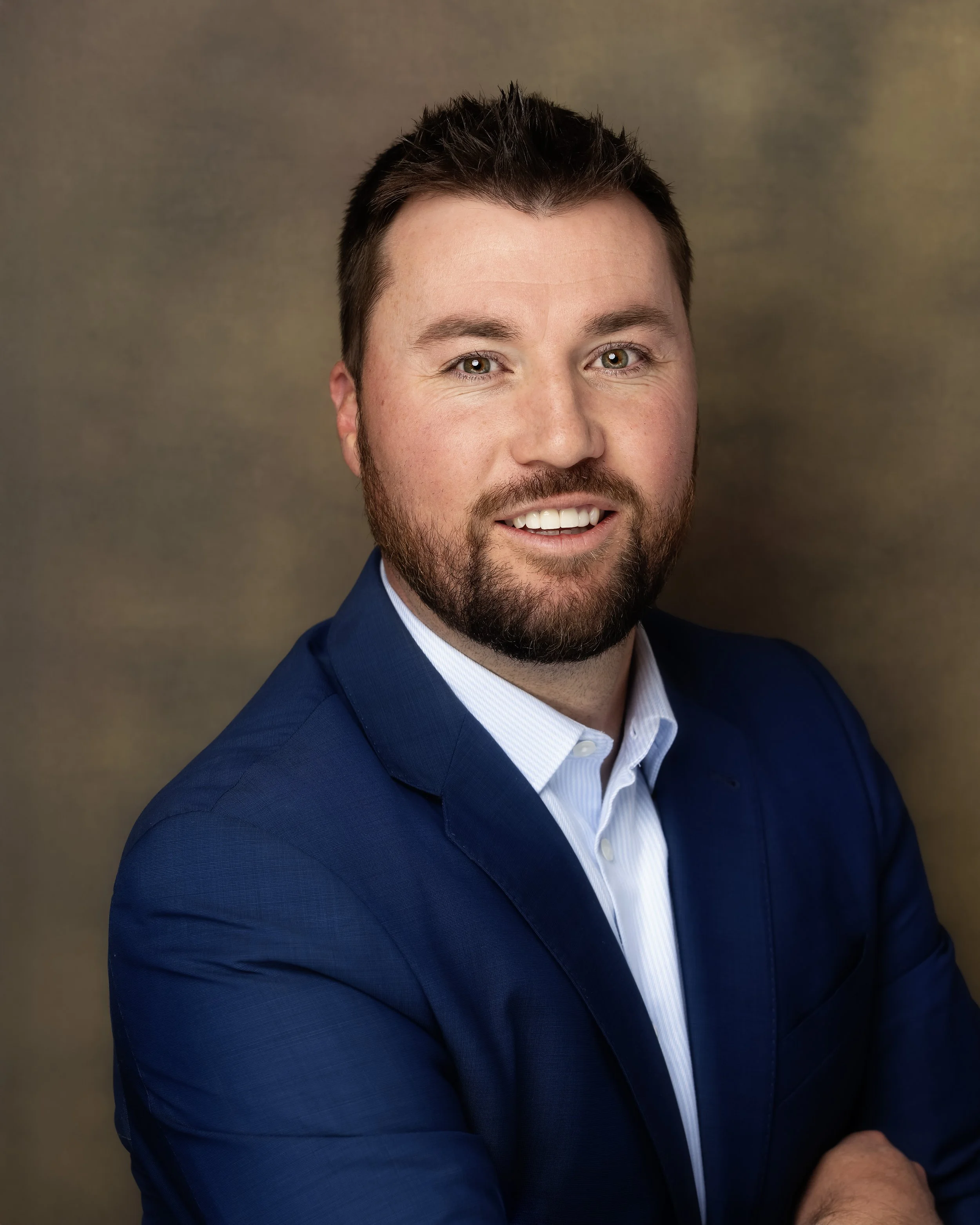 Professional headshot of a man with a beard and short dark hair, wearing a navy blazer and white shirt, smiling at the camera against a blurred brown background.