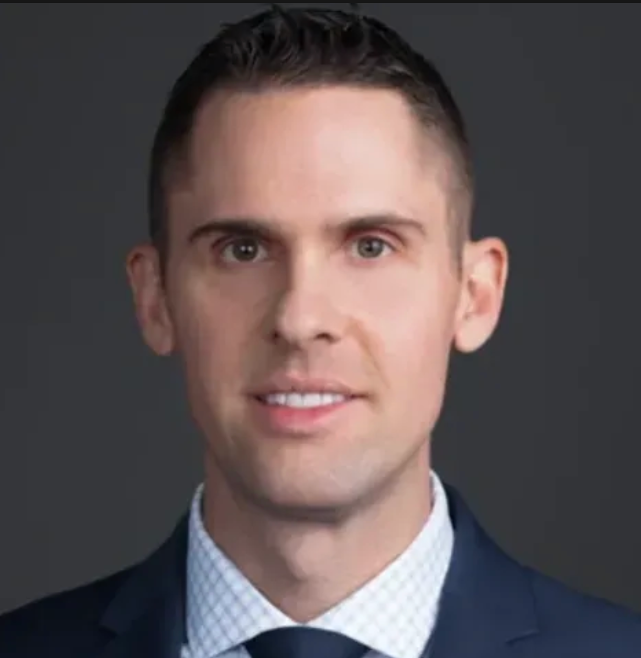Close-up portrait of a young man with short dark hair wearing a dark suit, white shirt, and black tie against a plain dark background.