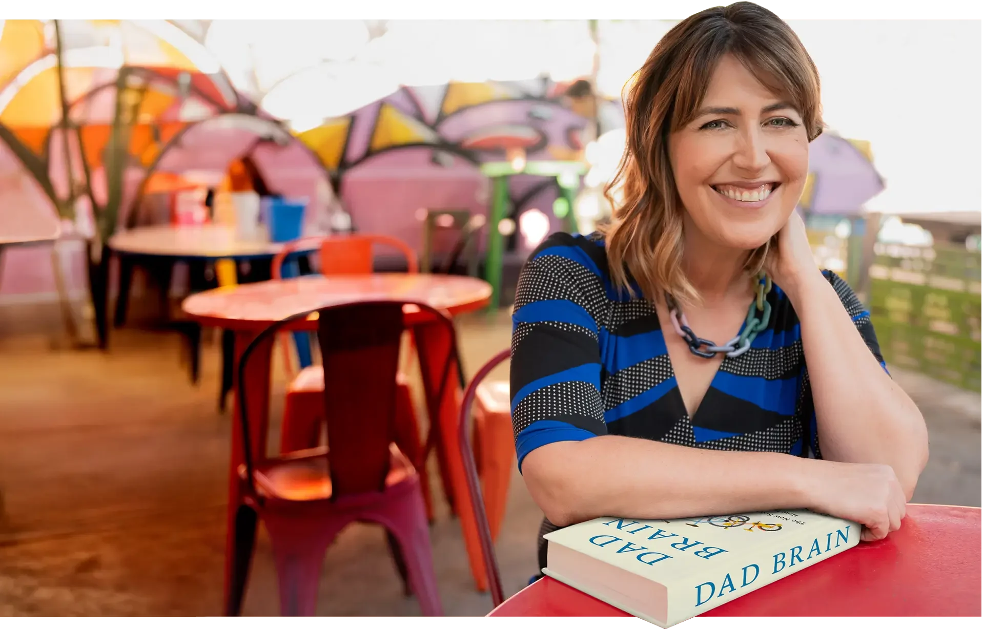 A woman sitting at an outdoor cafe table, smiling, with a large book titled 'Dad Brain' in front of her. The background shows colorful umbrellas and outdoor seating.