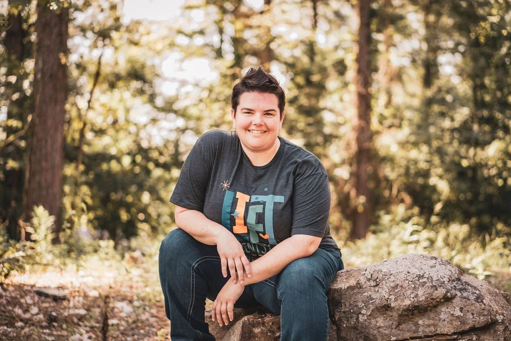 A young person sitting on a large rock in a forest, smiling at the camera, wearing a gray t-shirt and jeans, with trees and sunlight in the background.