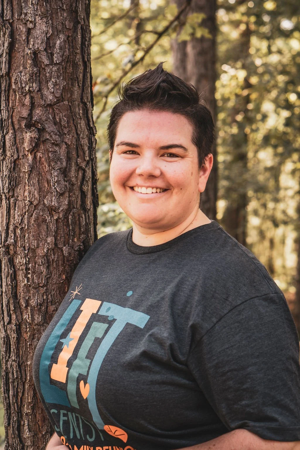 A person smiling outdoors next to a tree with a background of trees and sunlight.