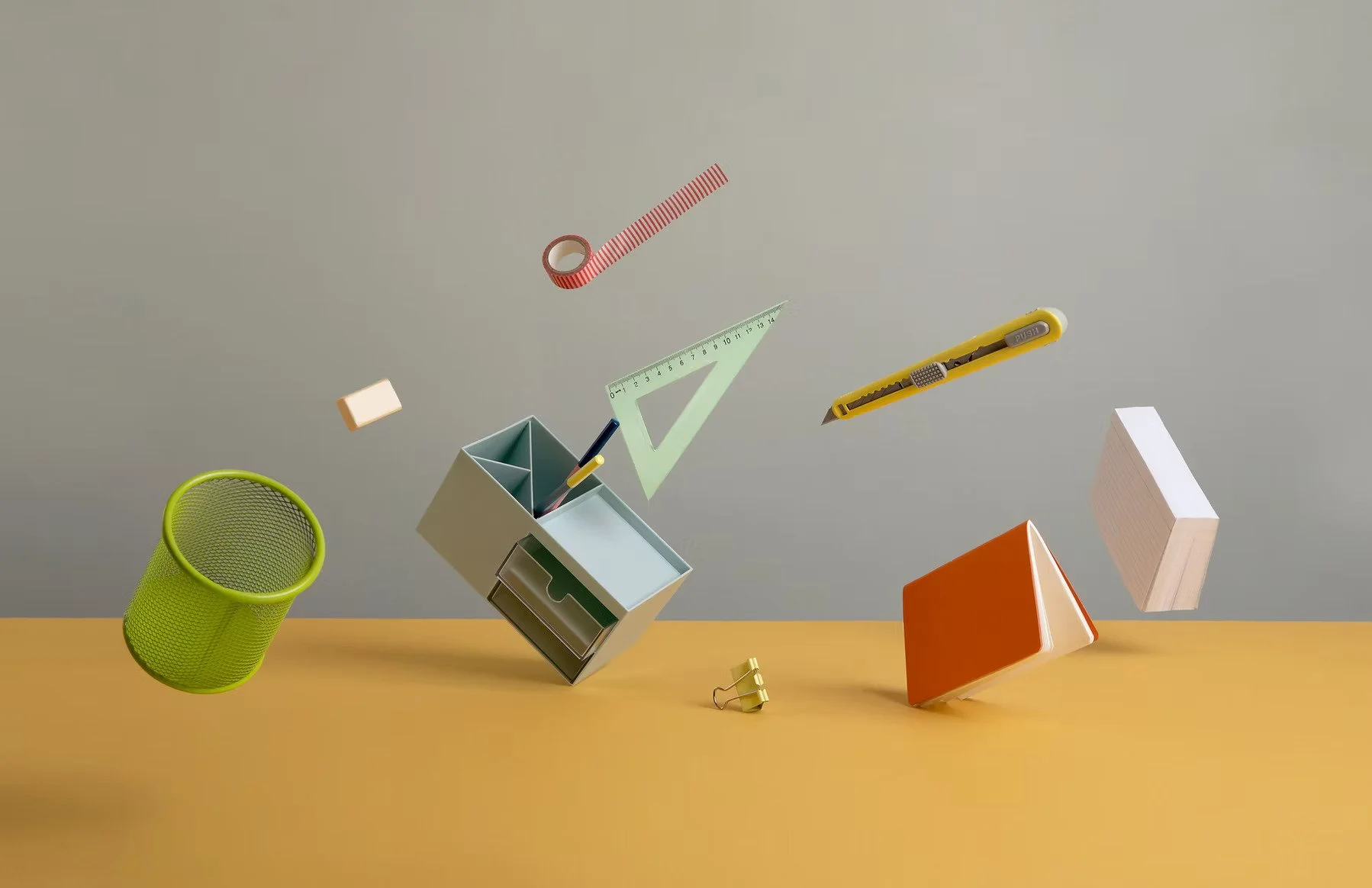 Floating office supplies including a green mesh pen holder, a small open desk organizer with pens, a yellow utility knife, a translucent green ruler, a small block of white chalk, a yellow binder clip, a red and white washi tape roll, and paper books, against a gray wall and yellow surface.
