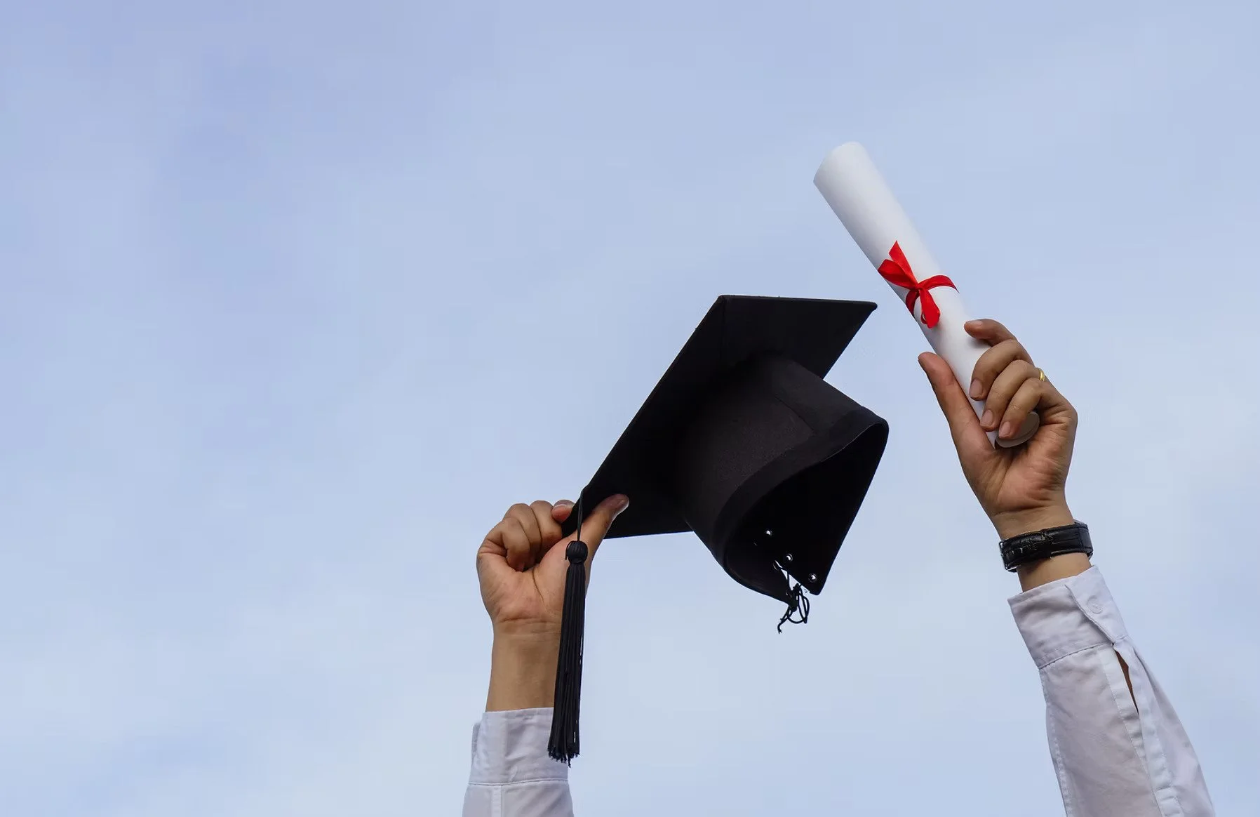 Person holding a graduation cap in one hand and a diploma in the other hand against a blue sky.